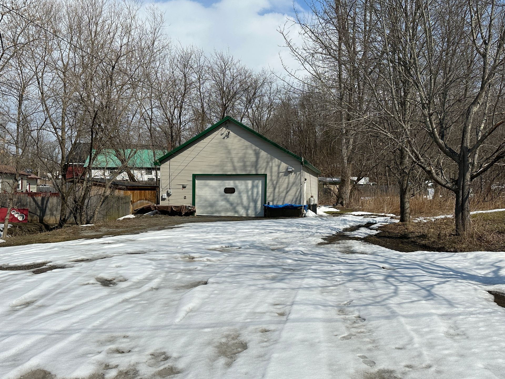 A detached garage with light-colored siding and a green roof sits at the end of a snowy, tire-tracked gravel driveway.