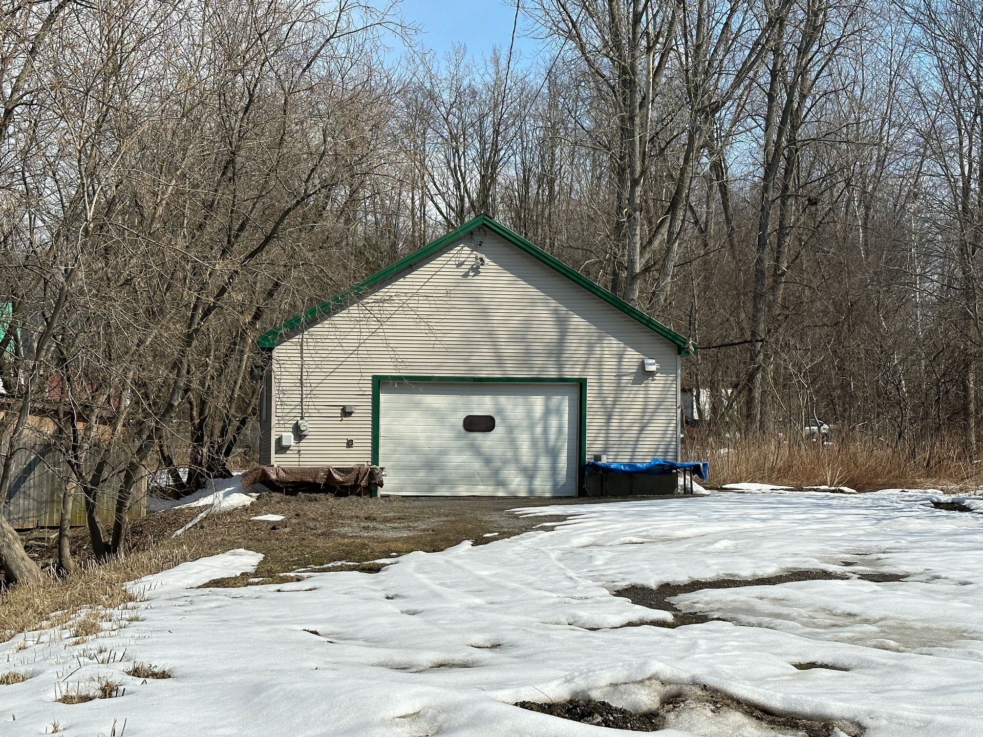 A beige, single-car garage with a green roof and trim, situated among bare trees with patches of snow on the ground.