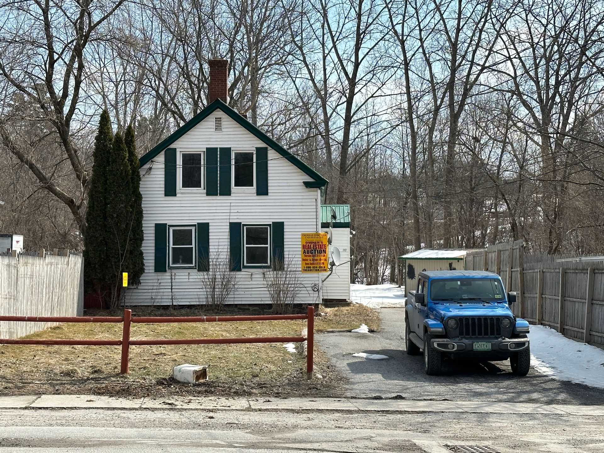 A white two-story house with green shutters and a brick chimney sits behind a wooden fence next to a parked blue pickup.