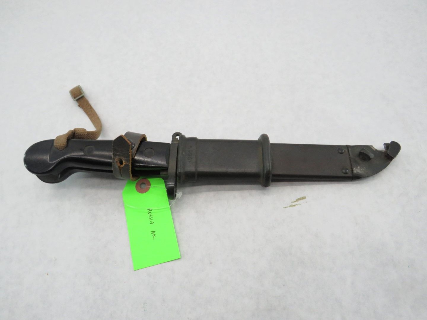 Black bayonet in a sheath with a leather strap and green tag.