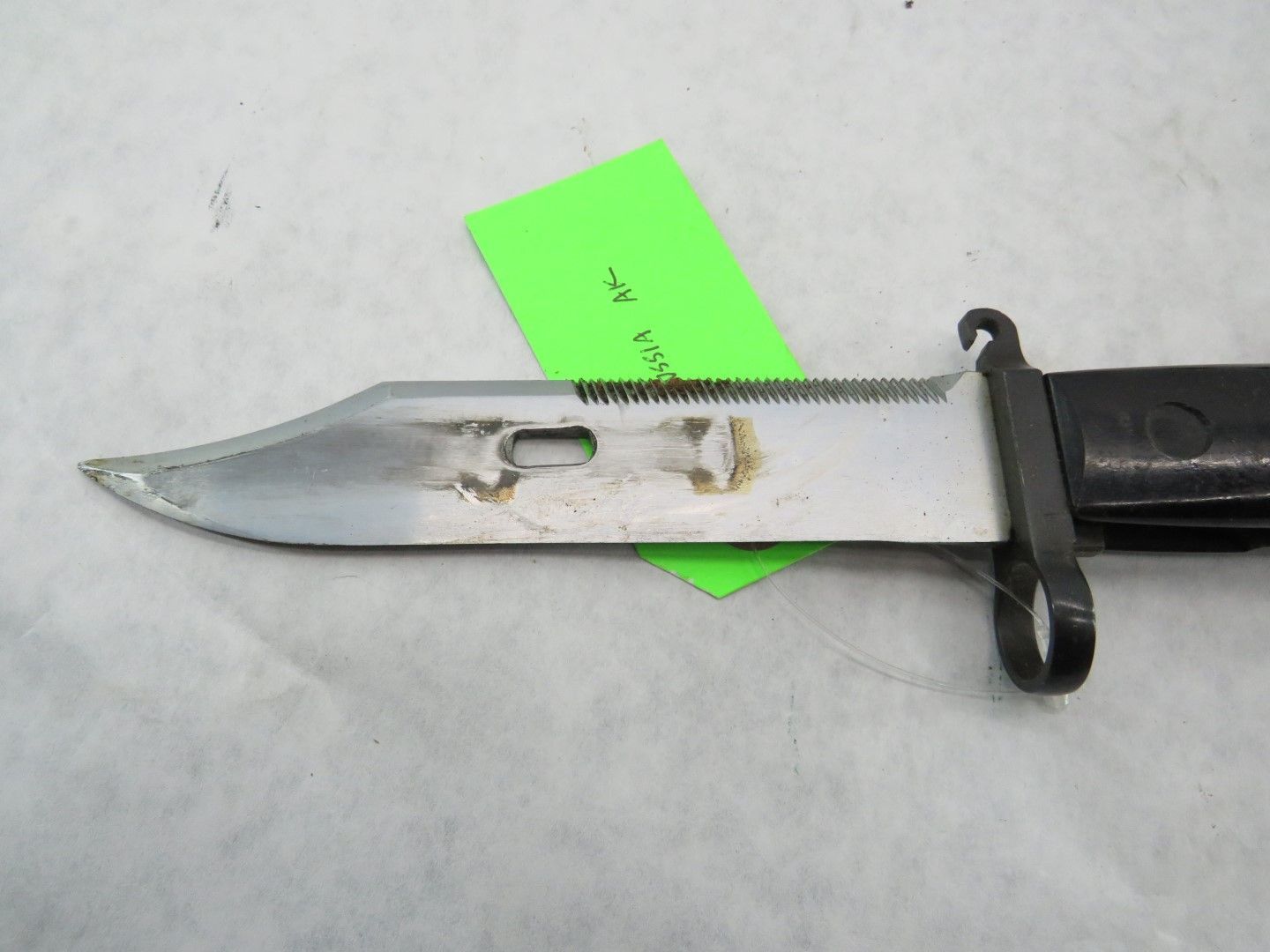 Bayonet with a silver blade and black handle, resting on a white surface. A green tag is attached to the blade.