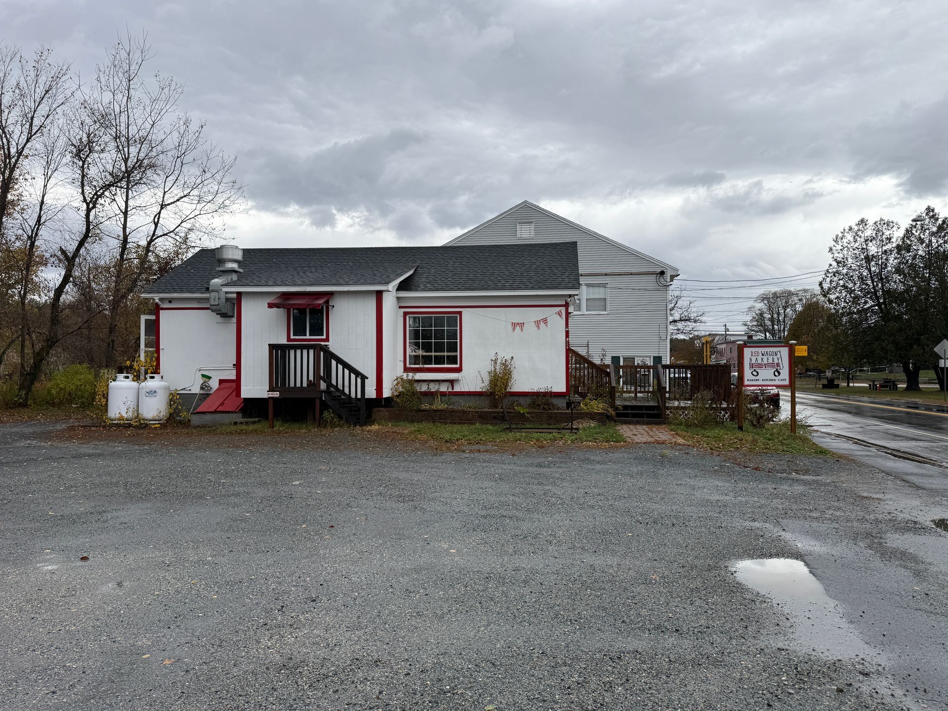 White building with red trim, gray siding, and a gravel parking lot under a cloudy sky.