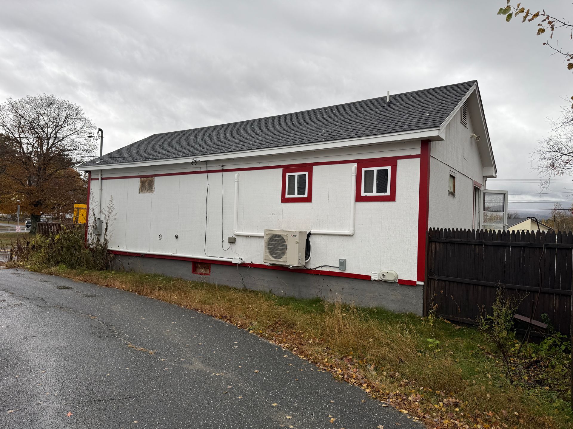 White building with red trim, gray roof, and two windows. An air conditioning unit is installed on the wall.