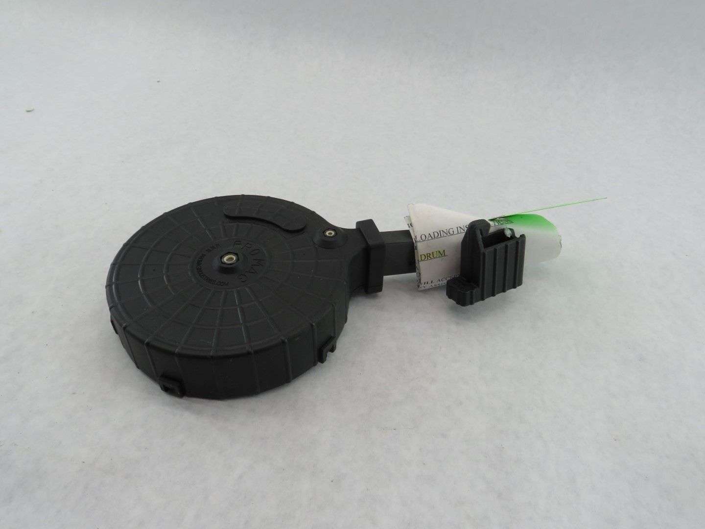 Black drum-style firearm magazine on a white surface, with a white and black attachment.