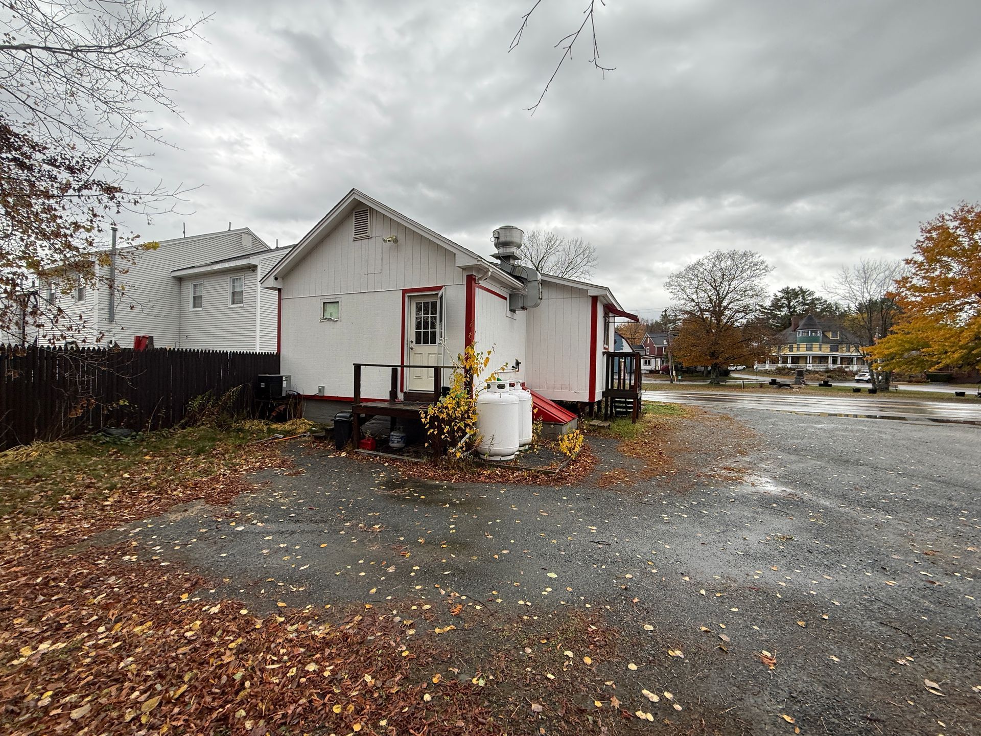 White building with red trim, parked propane tank, gravel lot, and autumn foliage under cloudy sky.