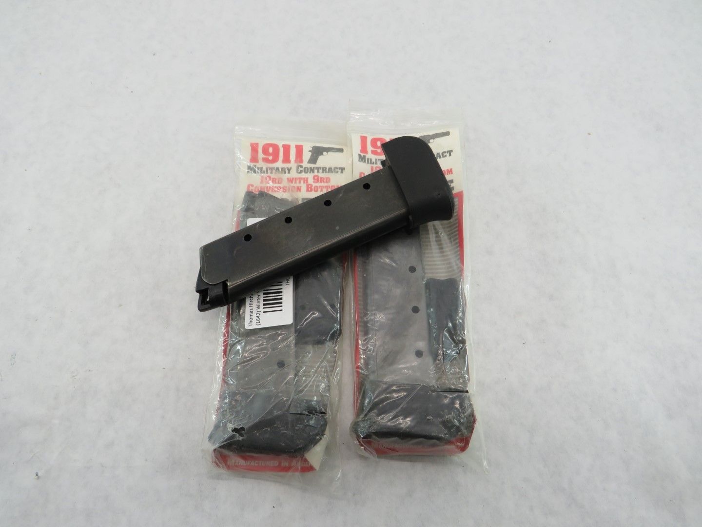 Two black 1911 pistol magazines, one loose, two sealed in packaging, on a white surface.
