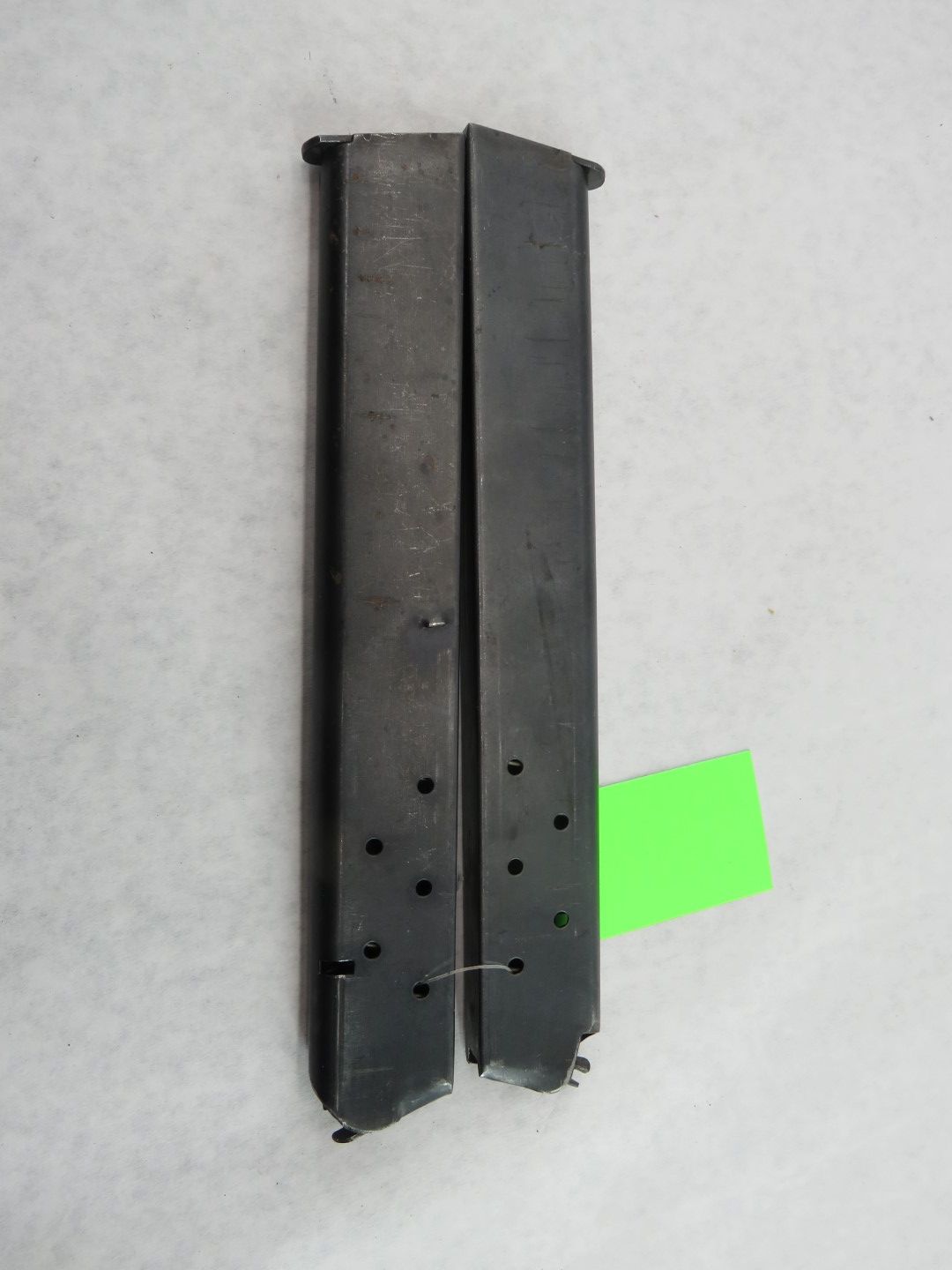 Two dark, metal handgun magazines with bullet indicator holes, against a white background. Green tag attached.