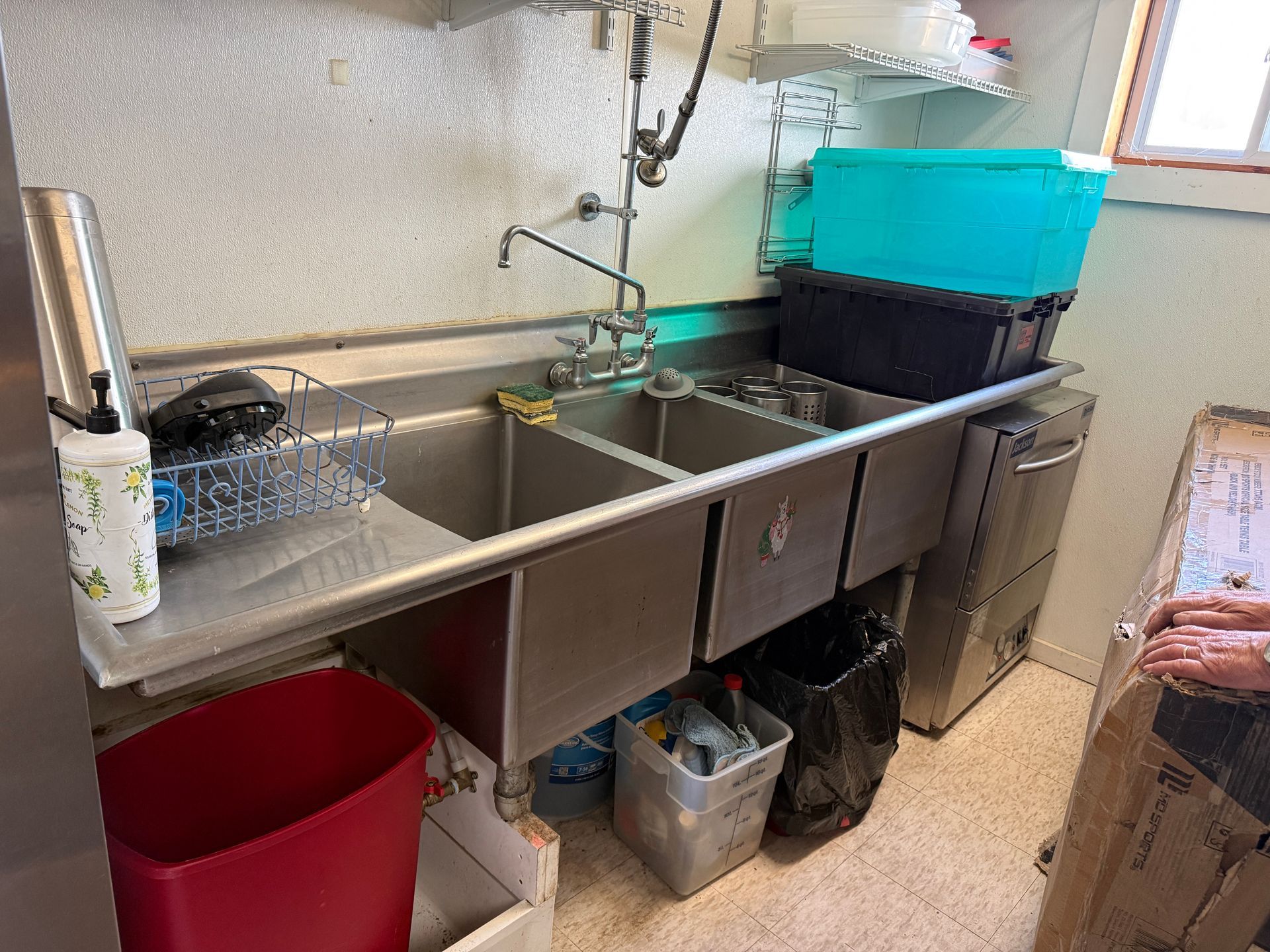 Stainless steel kitchen sink with three basins, overhead faucet, and nearby trash cans.