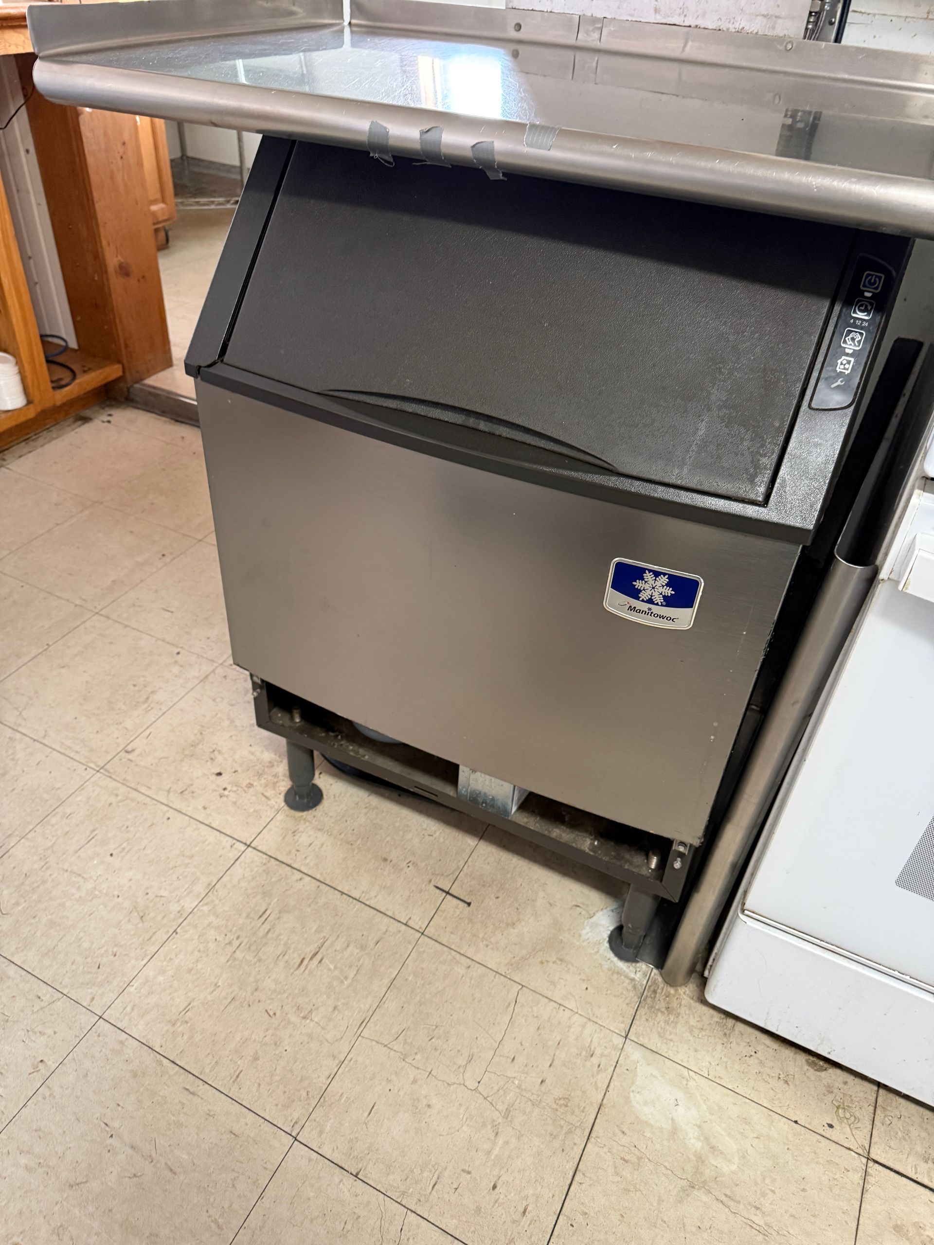 Stainless steel ice machine under a metal countertop in a commercial kitchen.