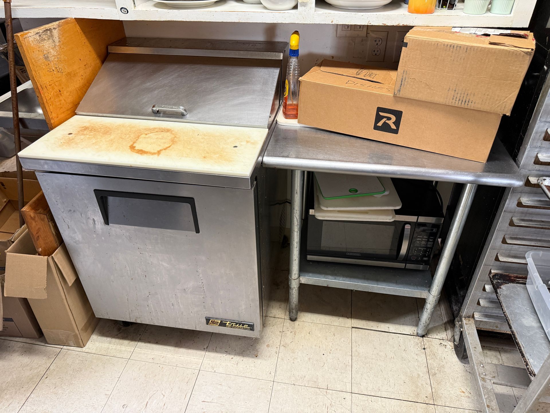 A commercial refrigerator with a cutting board top, next to a stainless steel table with boxes and supplies.