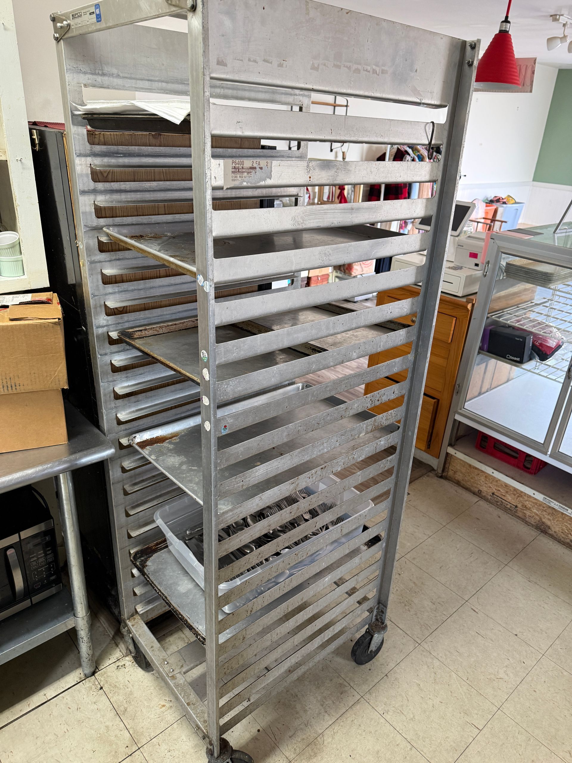 A tall, metal, wheeled rack with numerous horizontal shelves, possibly for baking trays, in a commercial kitchen setting.