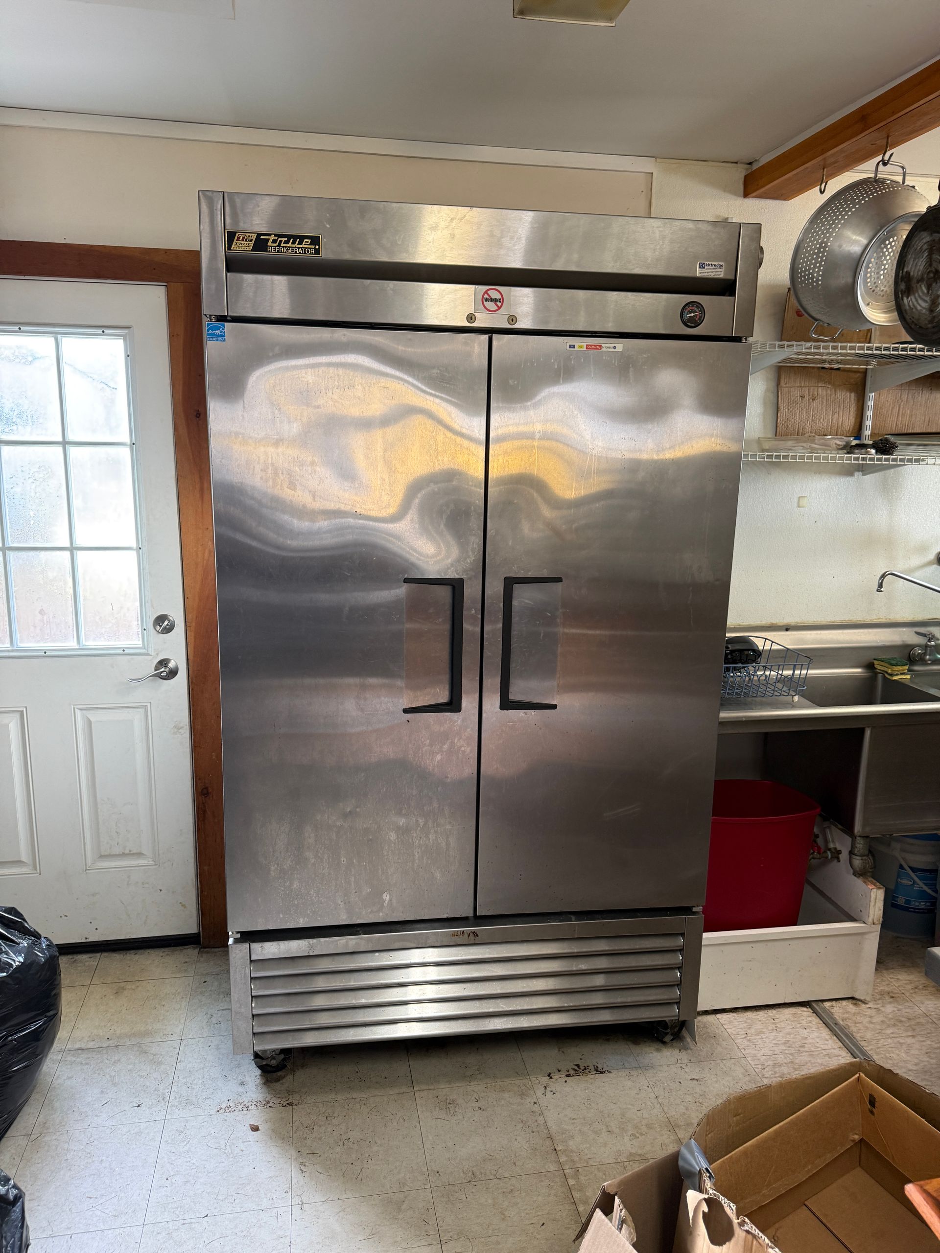 Stainless steel, double-door refrigerator in a kitchen setting; near a door, sink, and red bin.