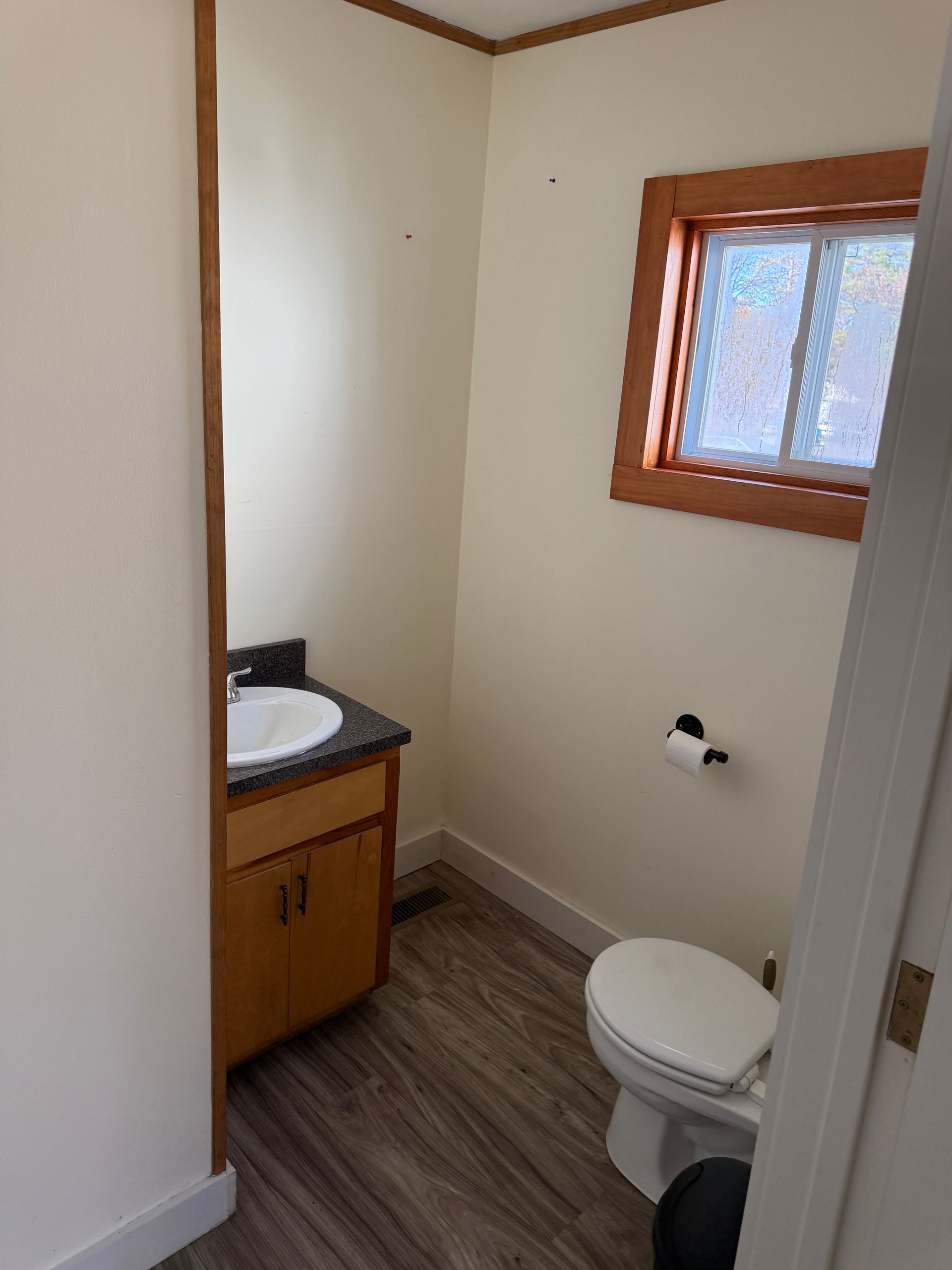 Small bathroom with toilet, sink, and window. Beige walls, wood trim, gray flooring.