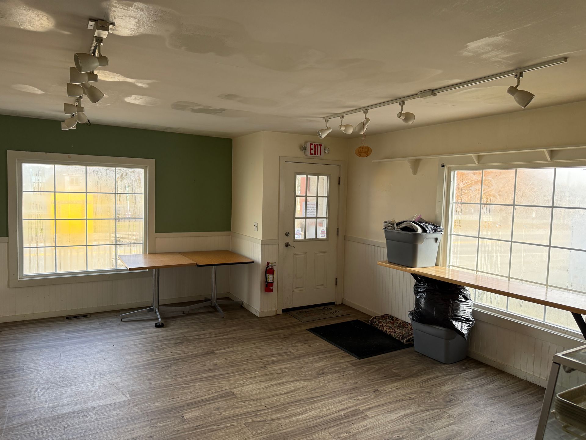 Empty room with two windows, a door, and track lighting. Tables line the walls.