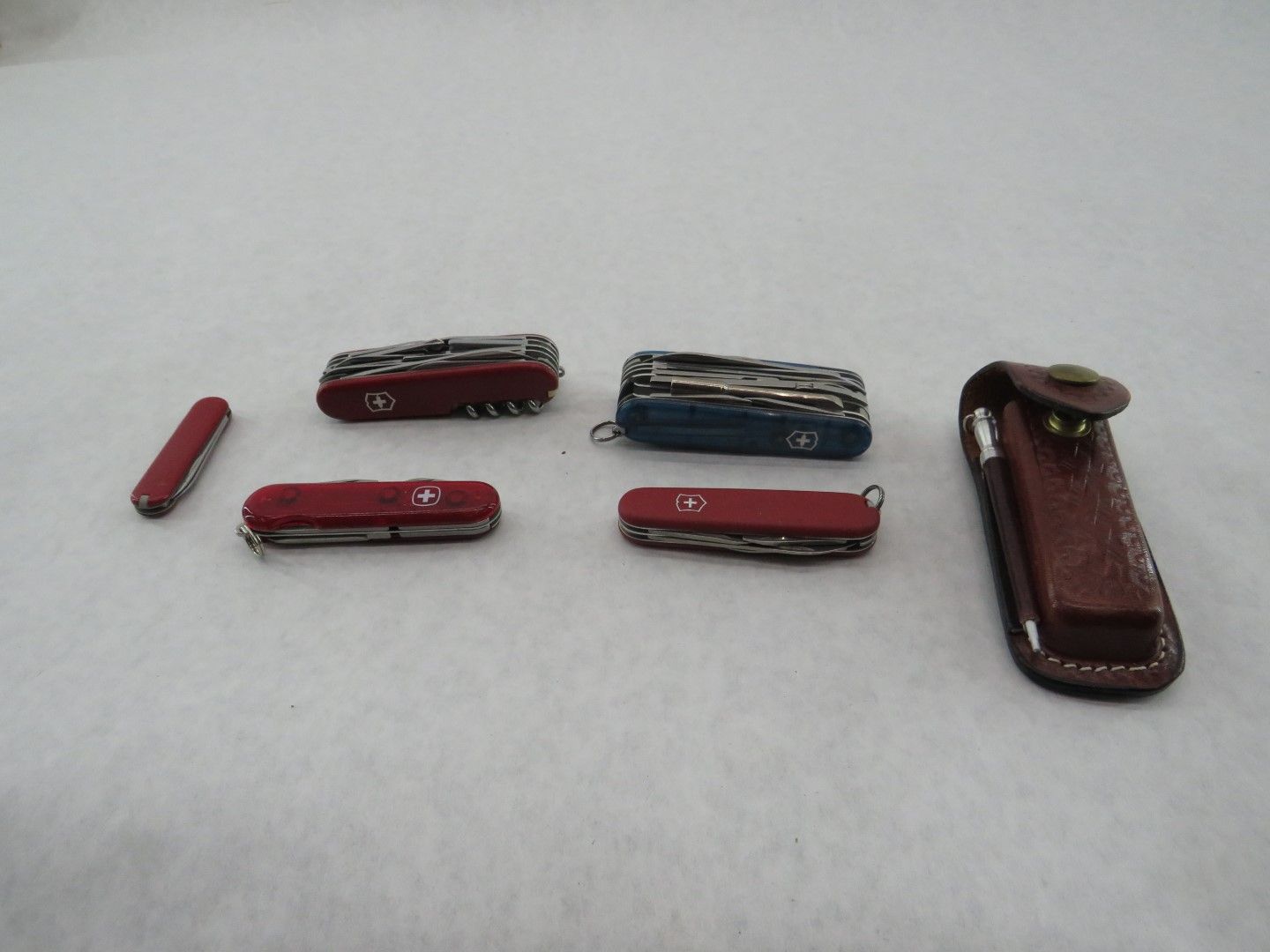 Several Swiss Army knives, red and blue, with a brown leather sheath, on a white surface.