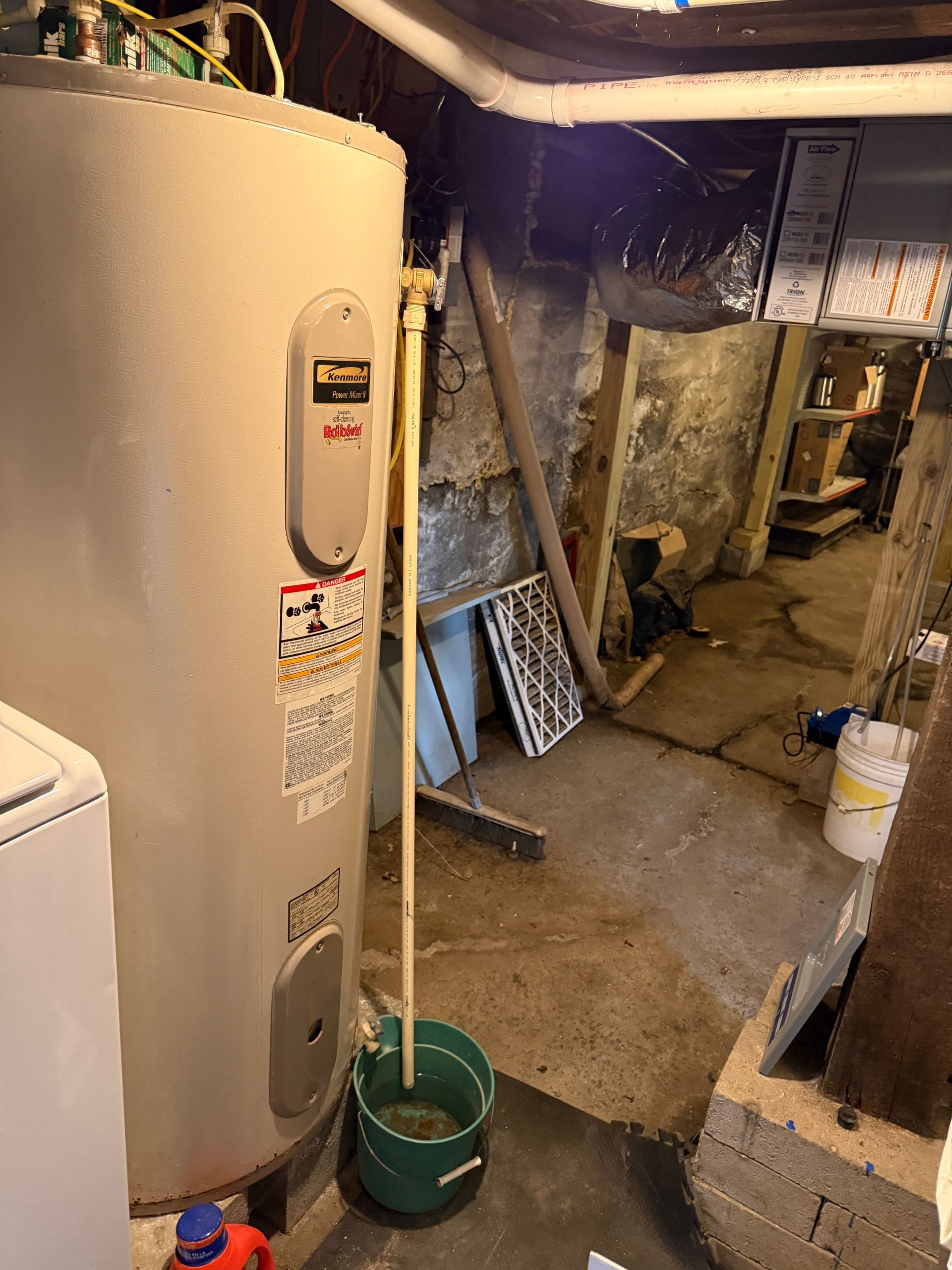 A water heater in a basement with a pipe draining into a bucket. Gray, concrete floor, stone wall.