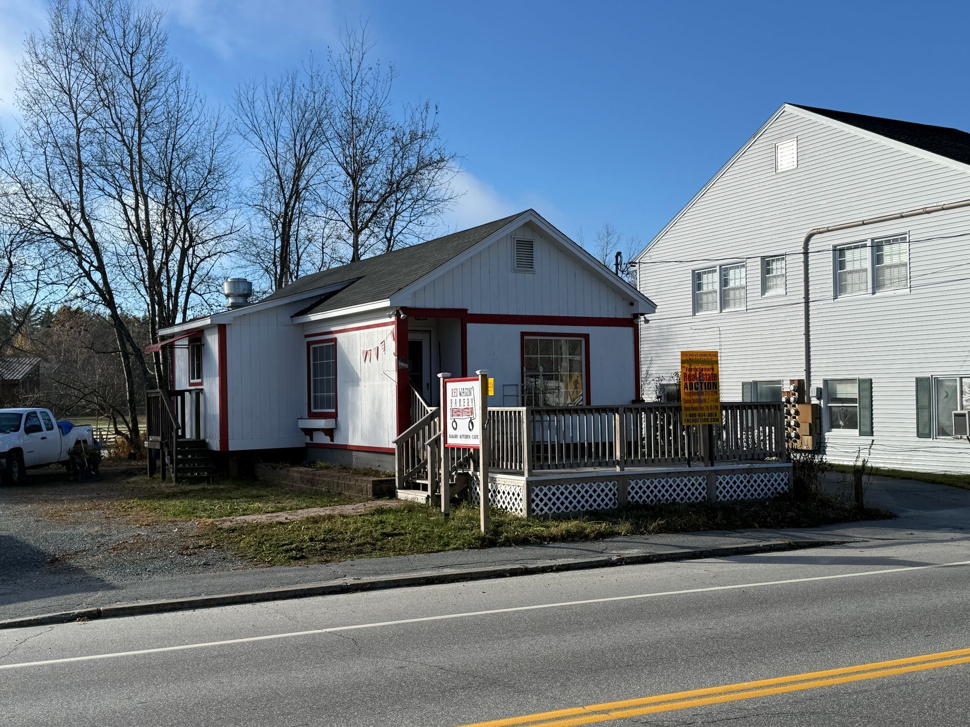 Small white building with red trim, deck, and sign, next to a larger white building, roadside.
