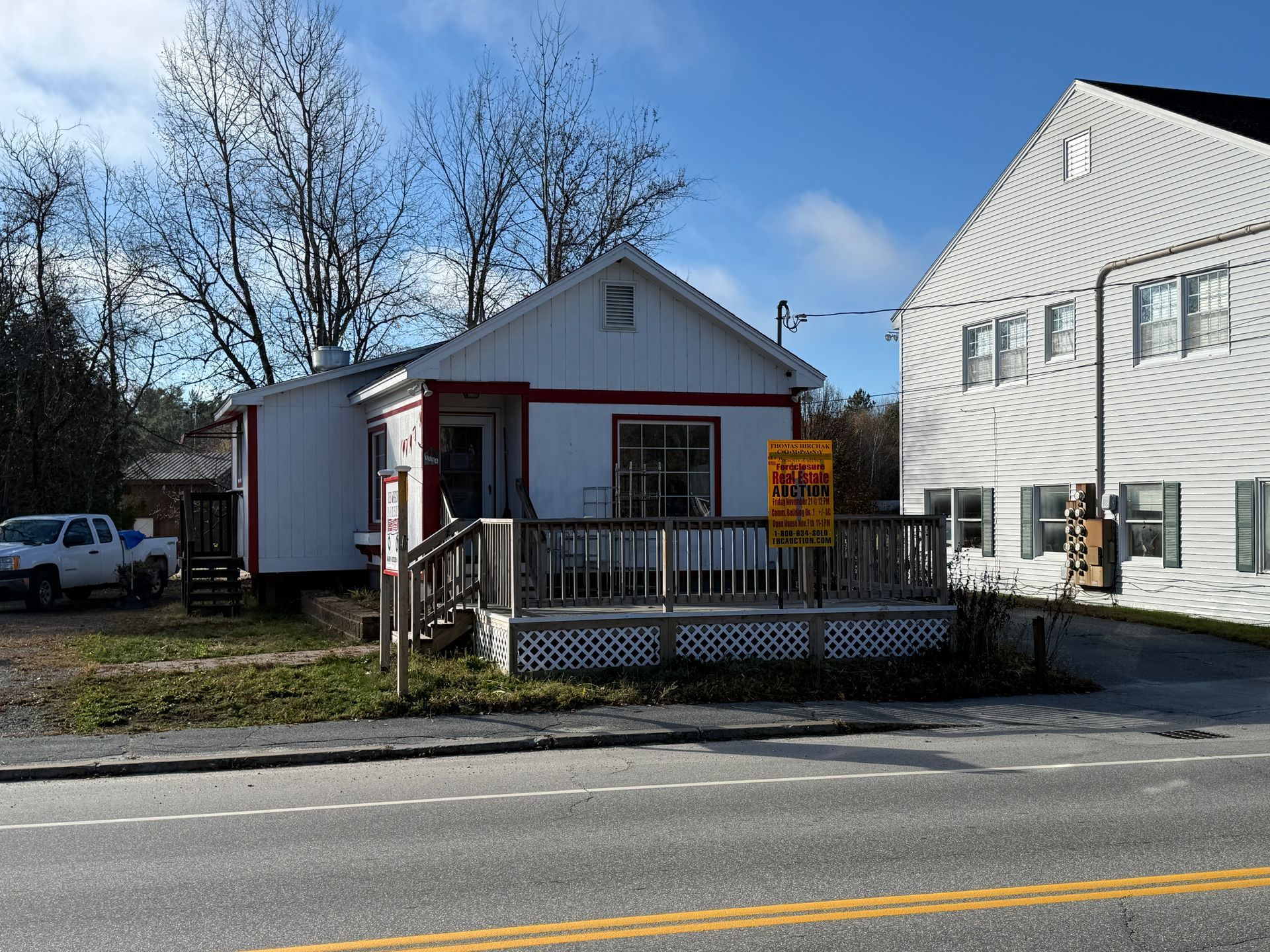 White building with red trim and a porch. Next to a larger white building and road.