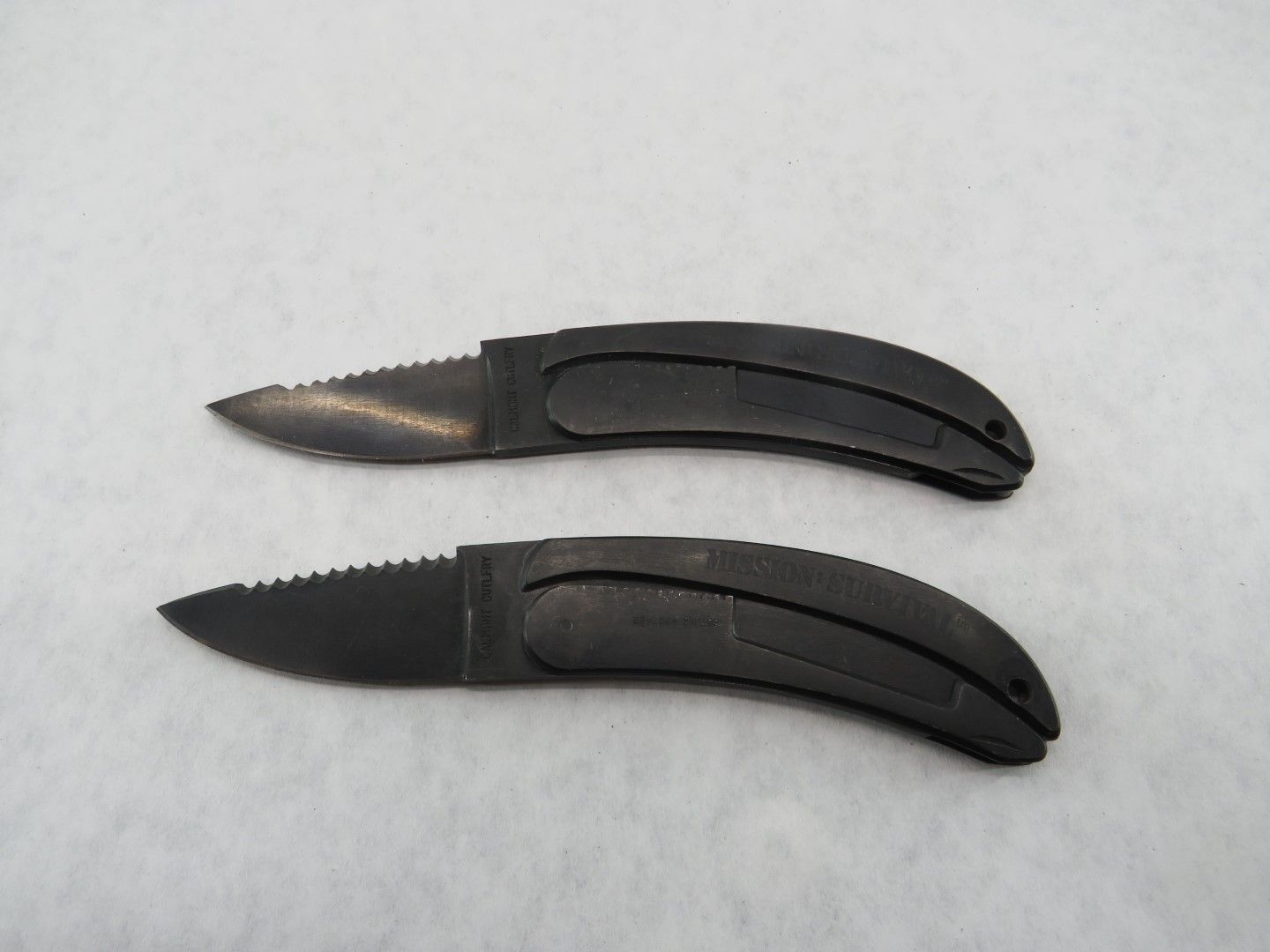 Two small black pocket knives with serrated blades, side by side, on a white surface.