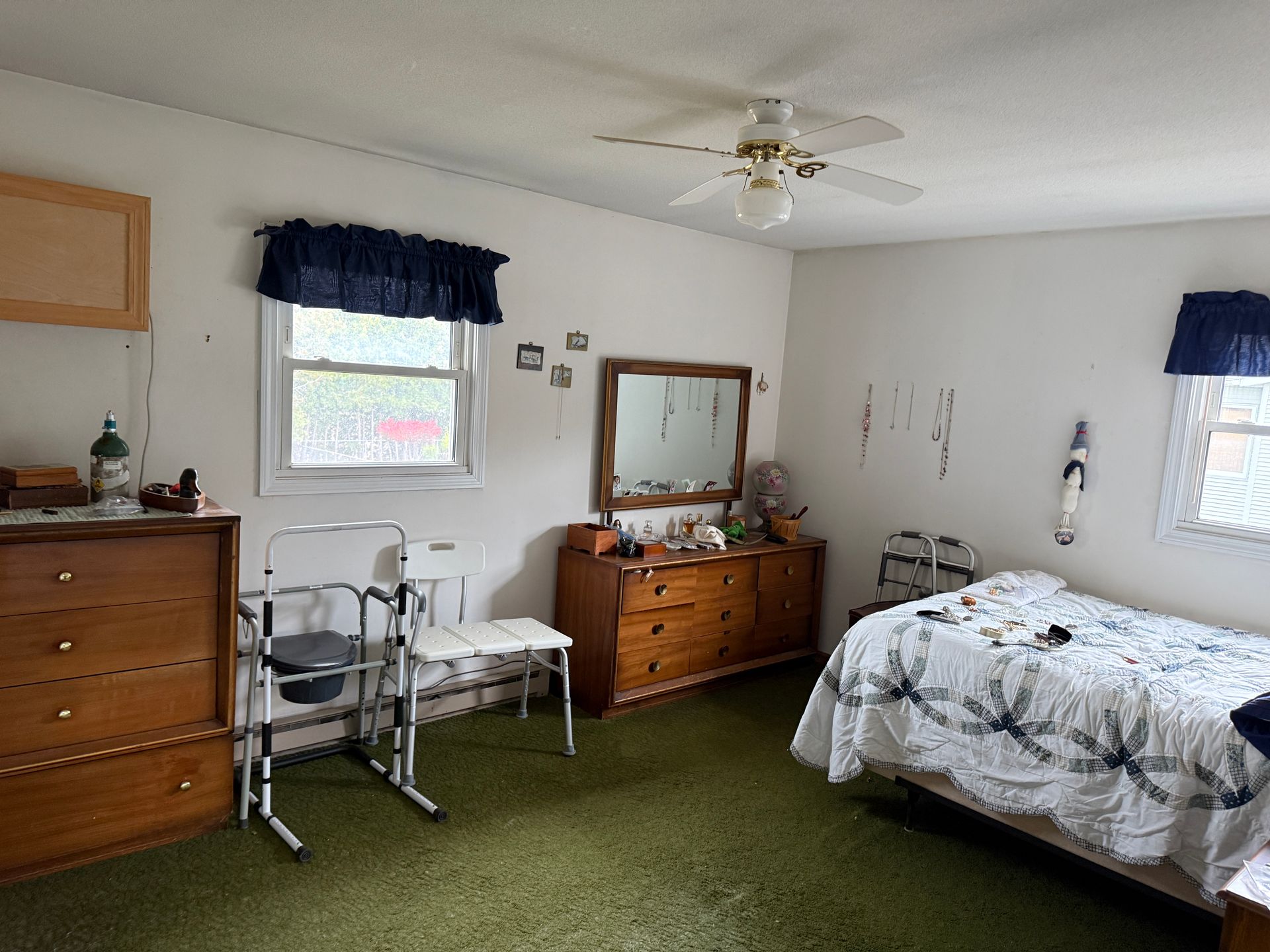 Bedroom with green carpet, wooden furniture, two windows, and medical equipment.