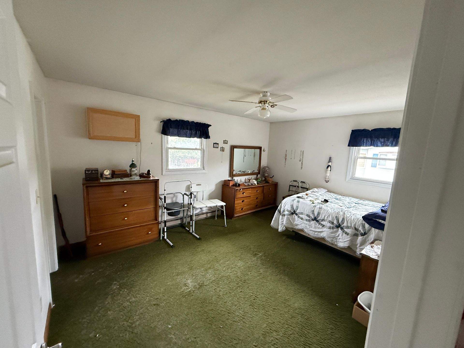 Bedroom with green carpet, wooden furniture, two windows with blue curtains, and a bed.