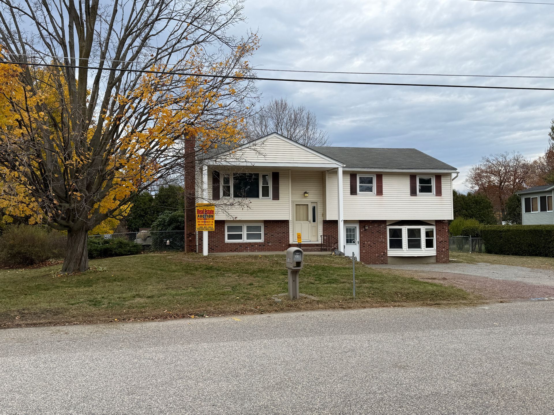 Two-story house with brick base, tan siding, brown shutters, and autumn tree in front yard.