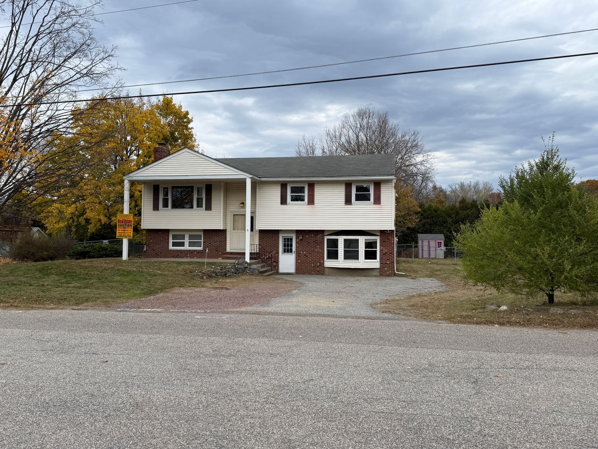 Beige two-story house with red brick and gravel driveway under a cloudy sky.