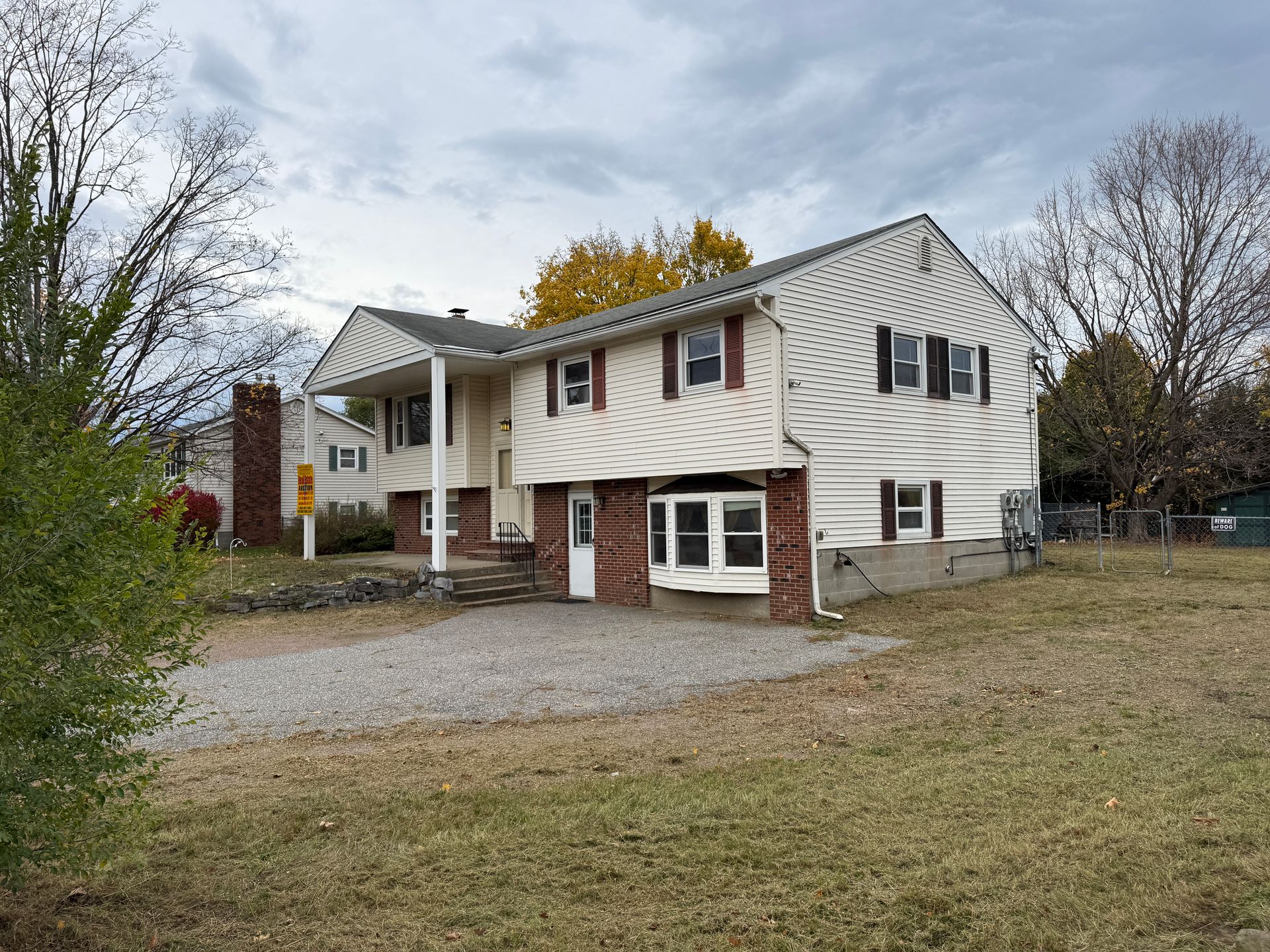 Two-story house with white siding and red shutters, a brick base, and a gravel driveway on a cloudy day.