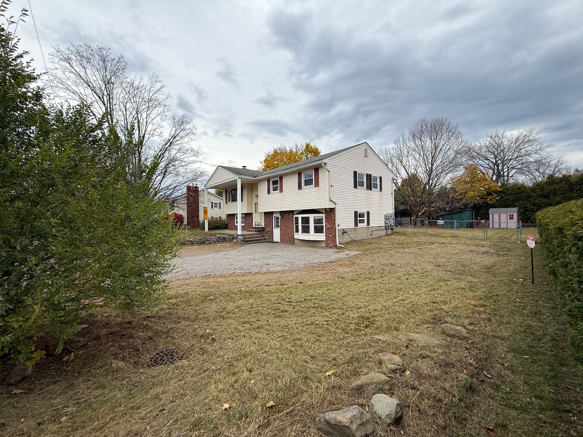 Two-story beige house with brick accents, overcast sky, lawn, gravel driveway, and some trees.