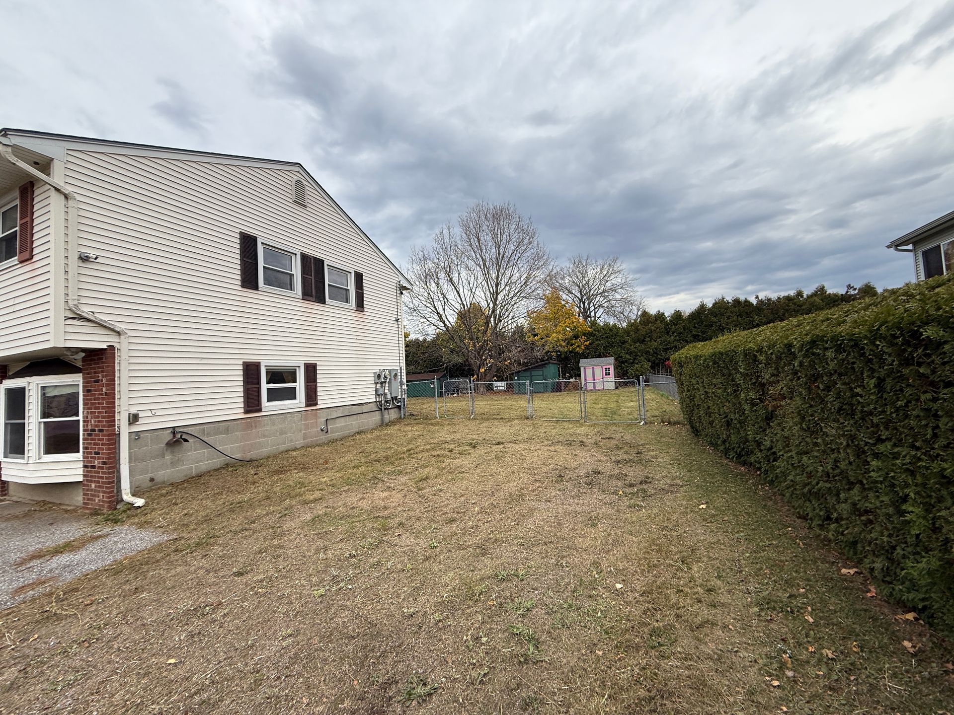 Side view of a house with a brown yard, a hedge, and cloudy sky.