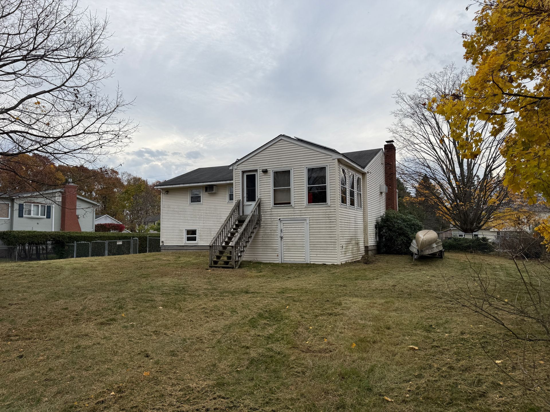 Back of a weathered, white house with a chimney and wooden steps; brown grass and overcast sky.