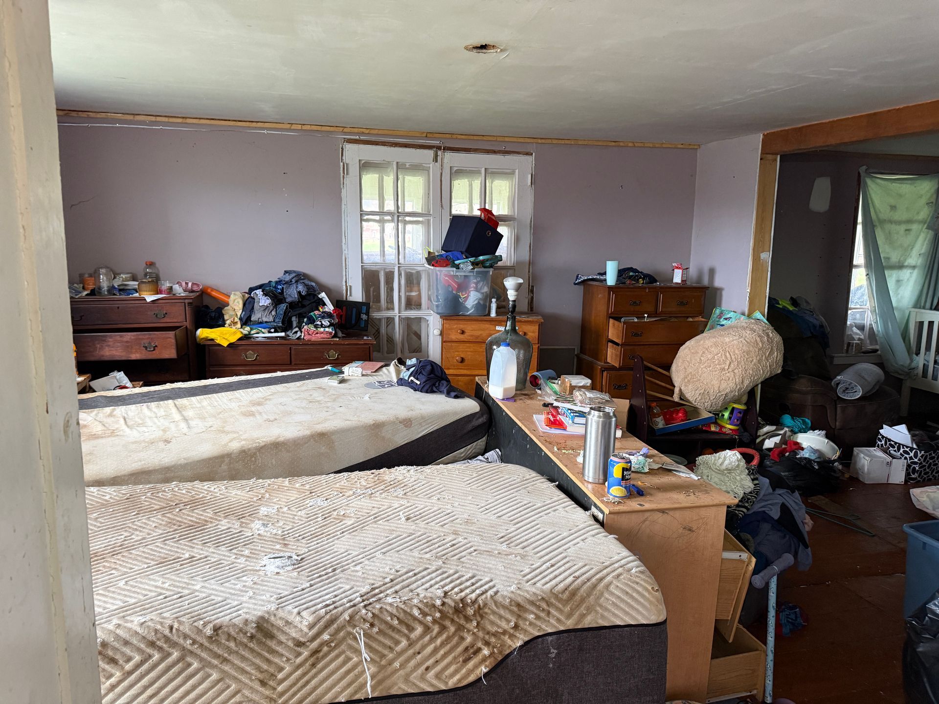 Bedroom with two beds, dressers, and various items. Light purple walls, window, and a doorway.