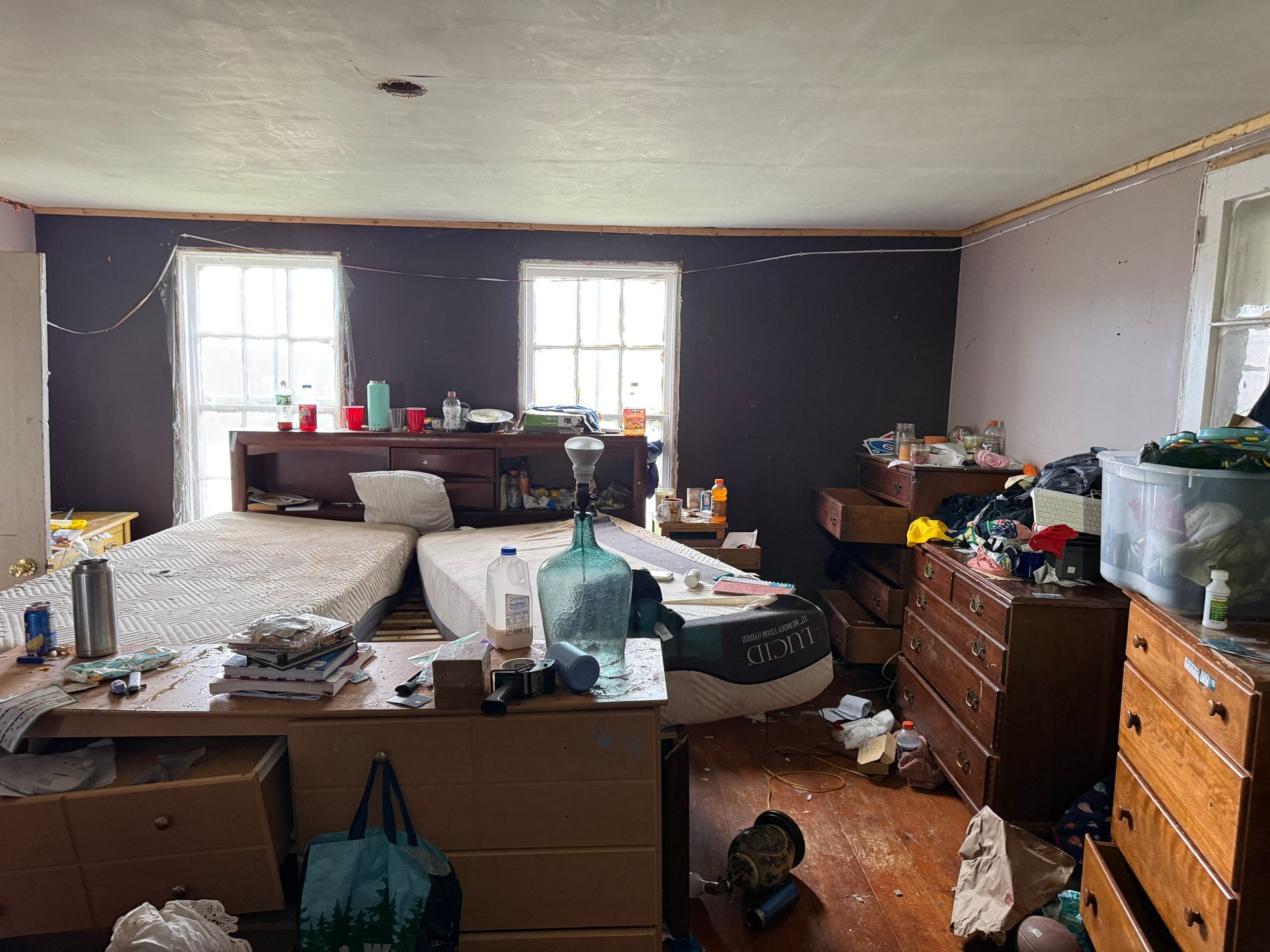 Bedroom: two beds, dressers with items, windows, purple wall, wooden floor.