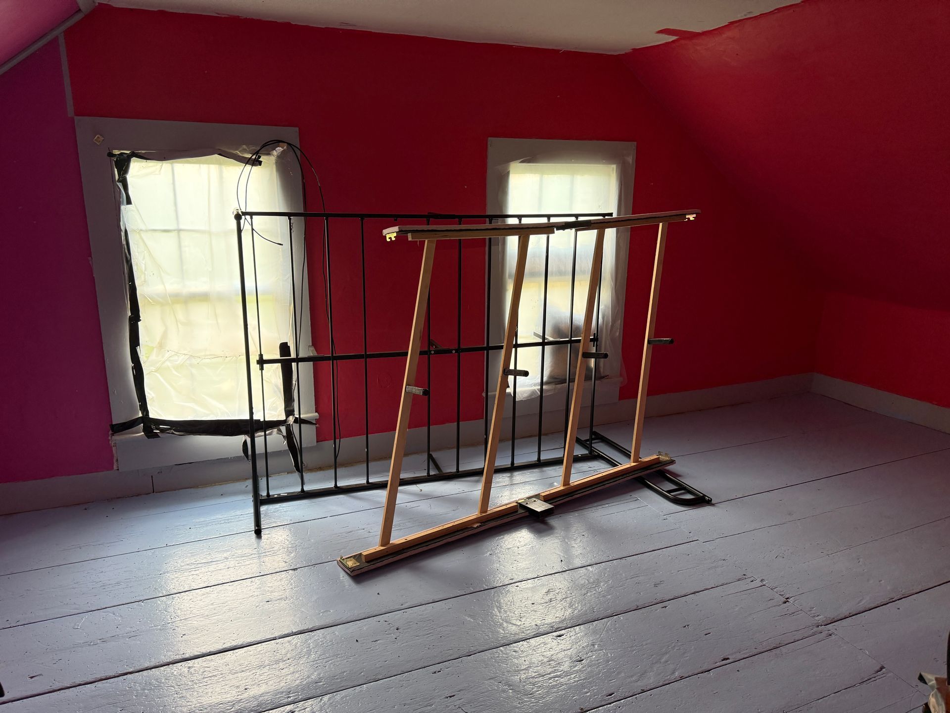 Red-walled room with two windows, painted white floor, metal bed frame.