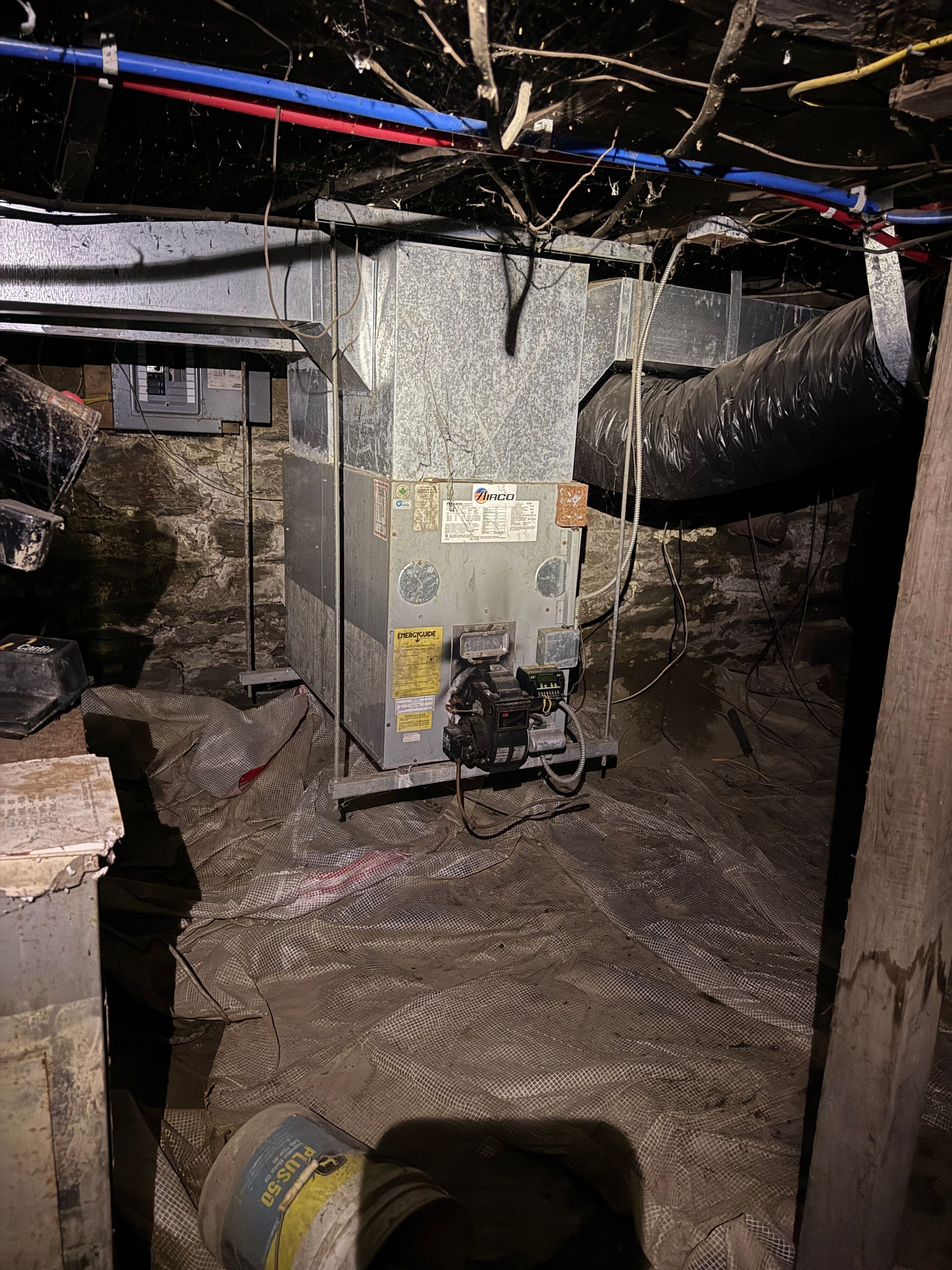 Furnace in a dirt-floored basement with pipes and ductwork overhead; gray and brown tones.