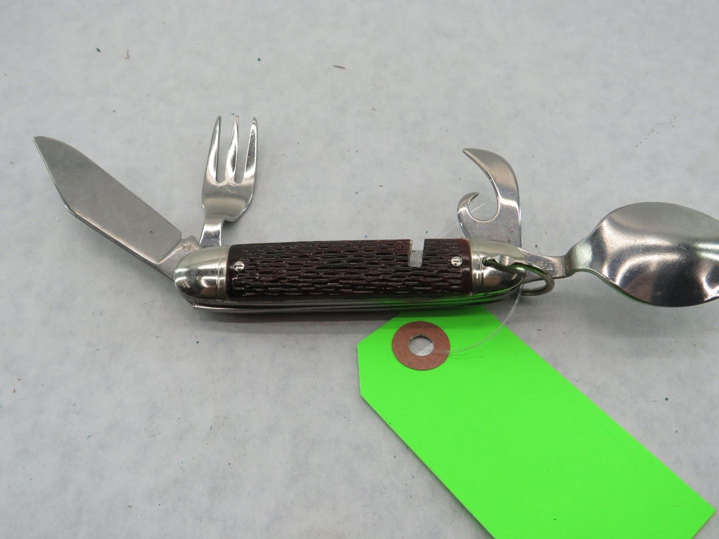 Multi-tool with knife, fork, can opener, and spoon, on a light surface with a green tag.