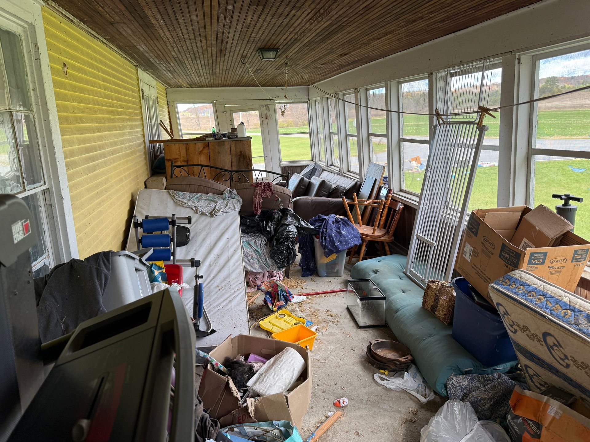 Sunroom filled with furniture, boxes, and various items. Yellow brick wall, windows, and a view of a field.