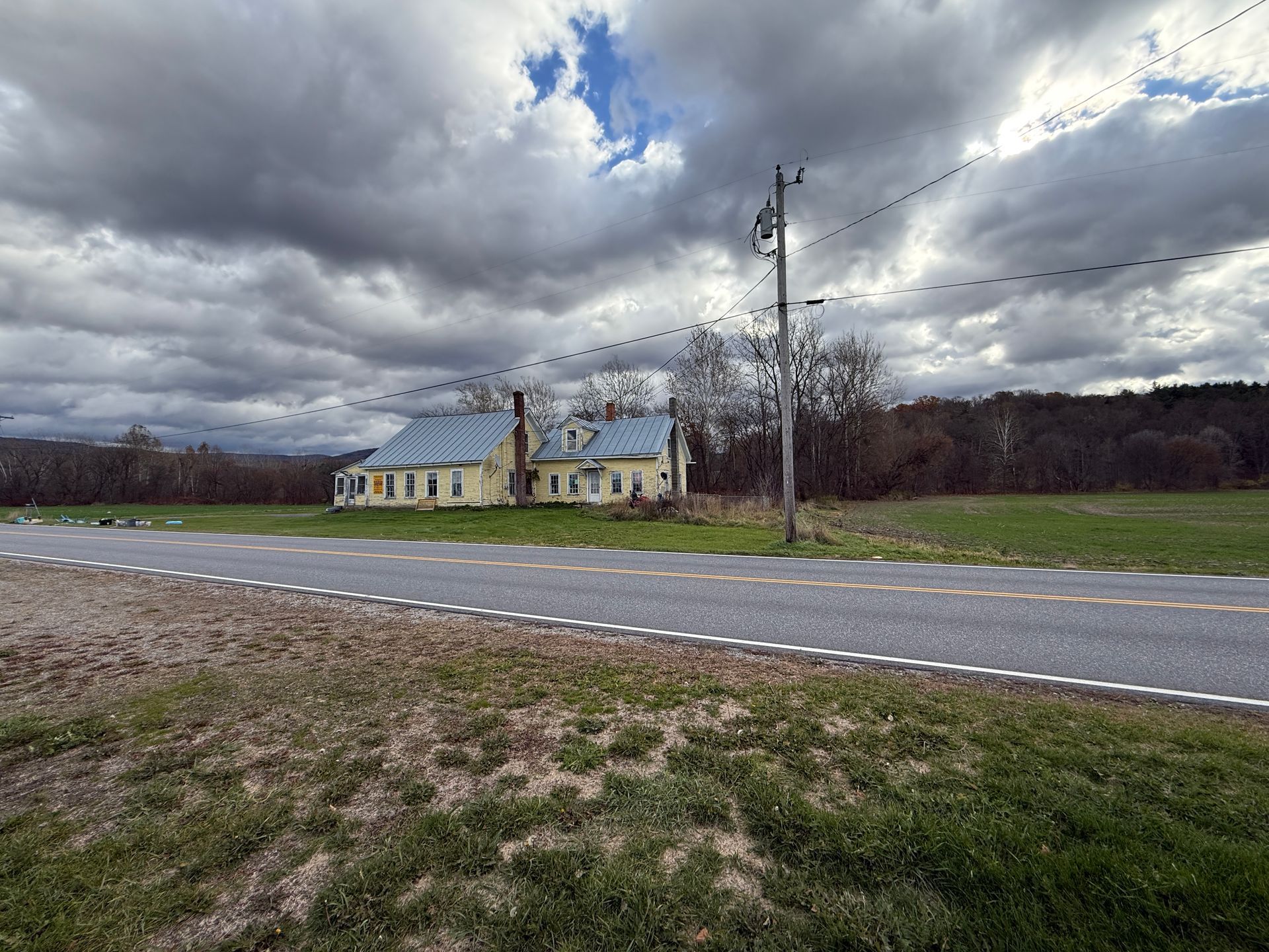 Yellow house with grey roof and overcast sky; road and grassy field in foreground.