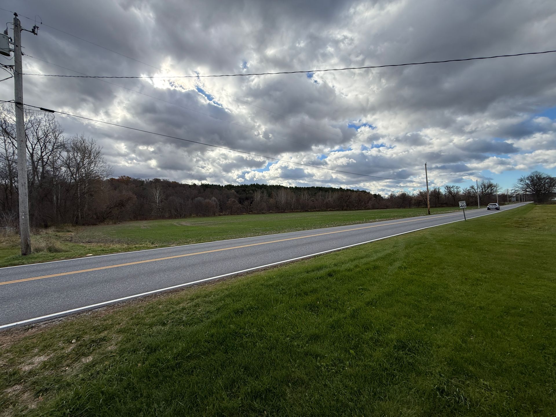 Road through a grassy field under a cloudy sky. Power lines are visible overhead.