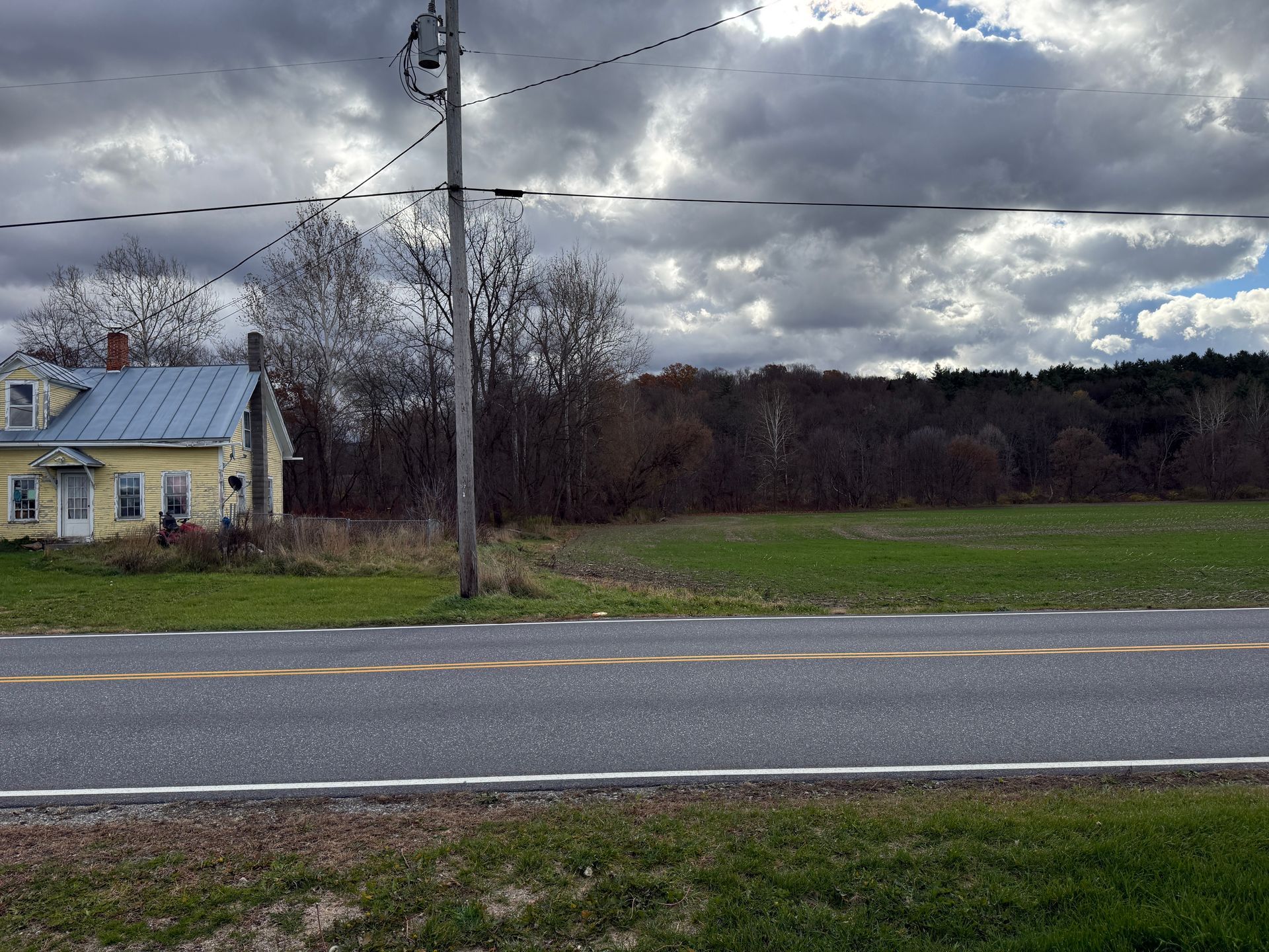 Yellow house beside a field with a road in front. Power lines overhead with a cloudy sky.
