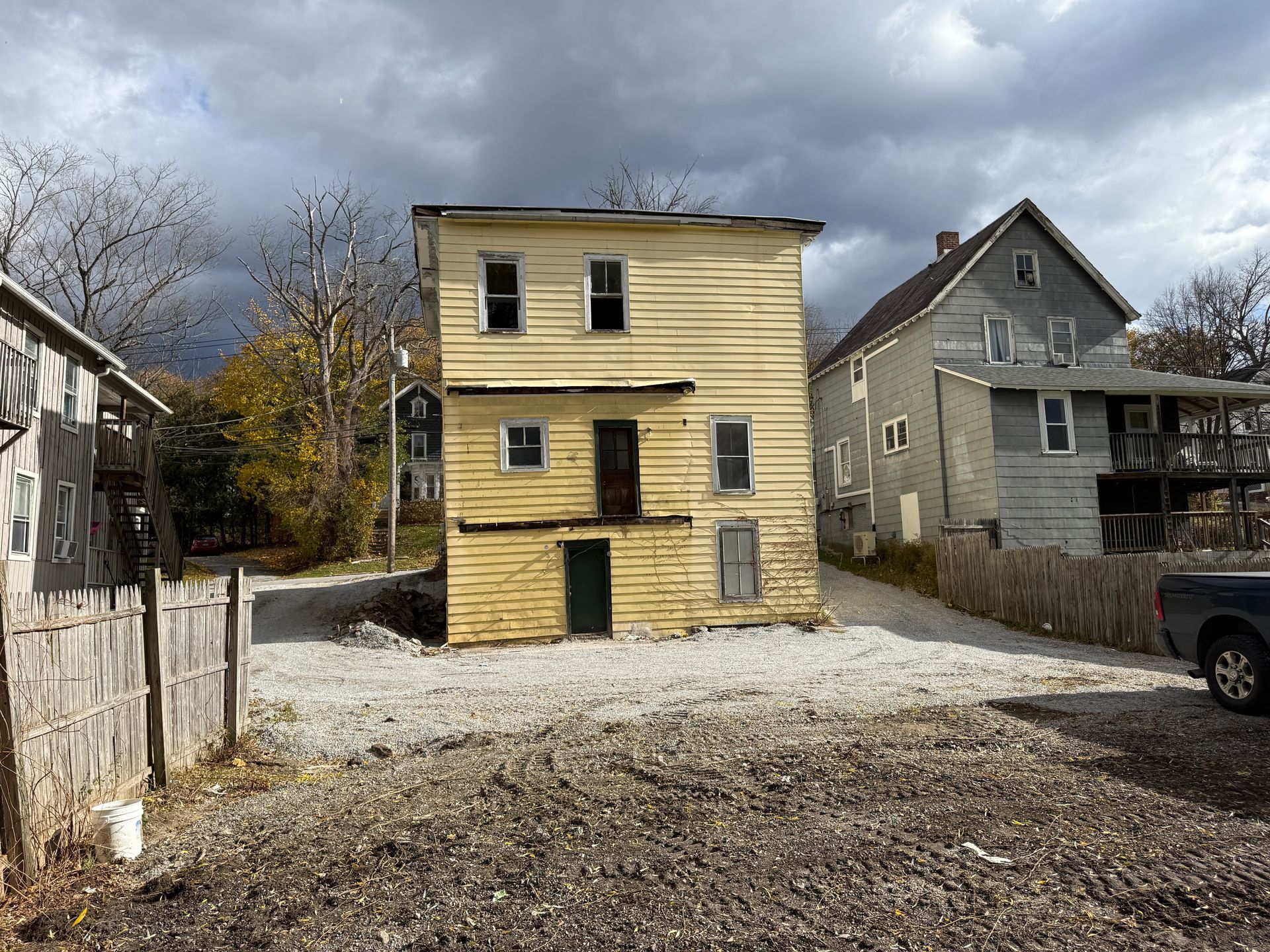 Yellow two-story building between two other structures on a gravel lot under a cloudy sky.