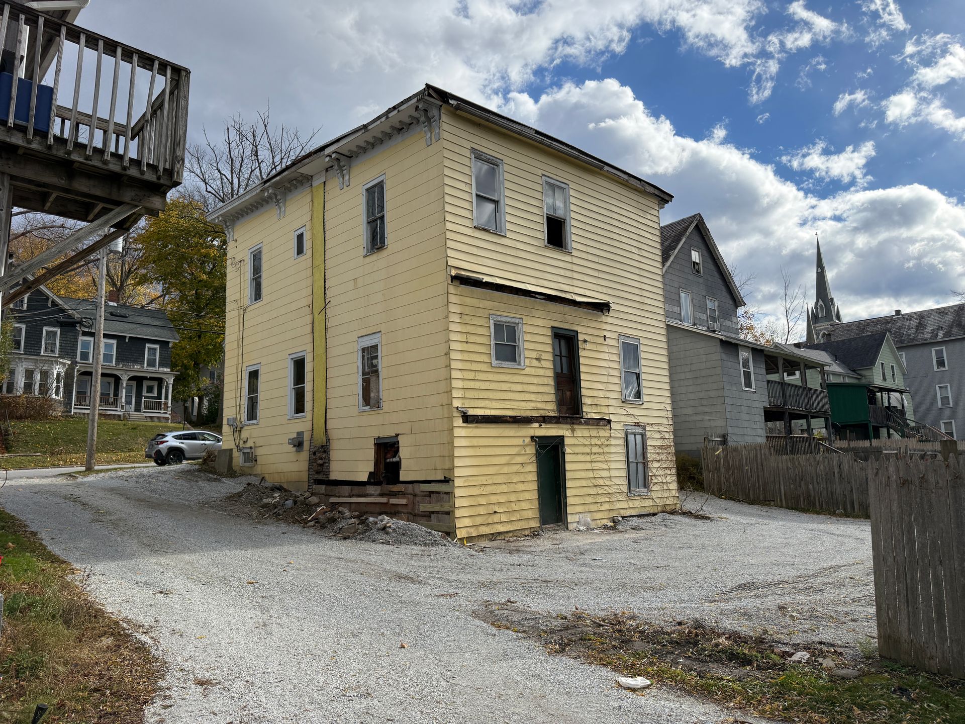 Yellow multi-story house with missing siding, gravel lot, and a church steeple in the distance.