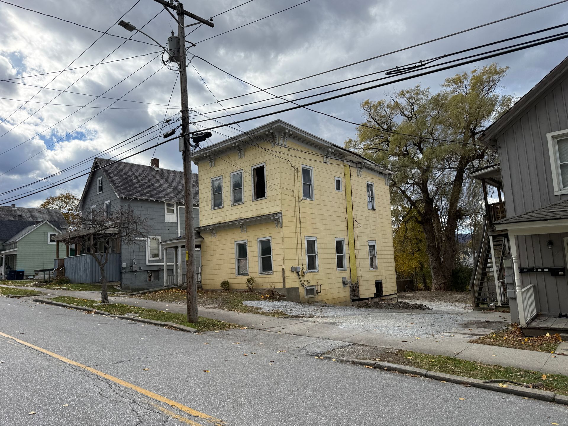 Yellow two-story house, gray sky, utility pole in front, other houses visible. Street in the foreground.