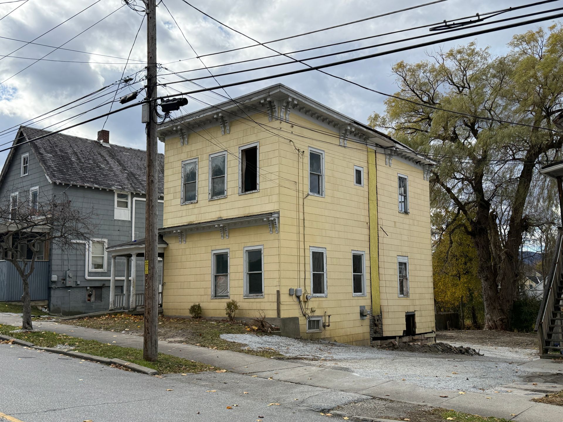 Two-story yellow house next to a gray house, both on a street under a cloudy sky.