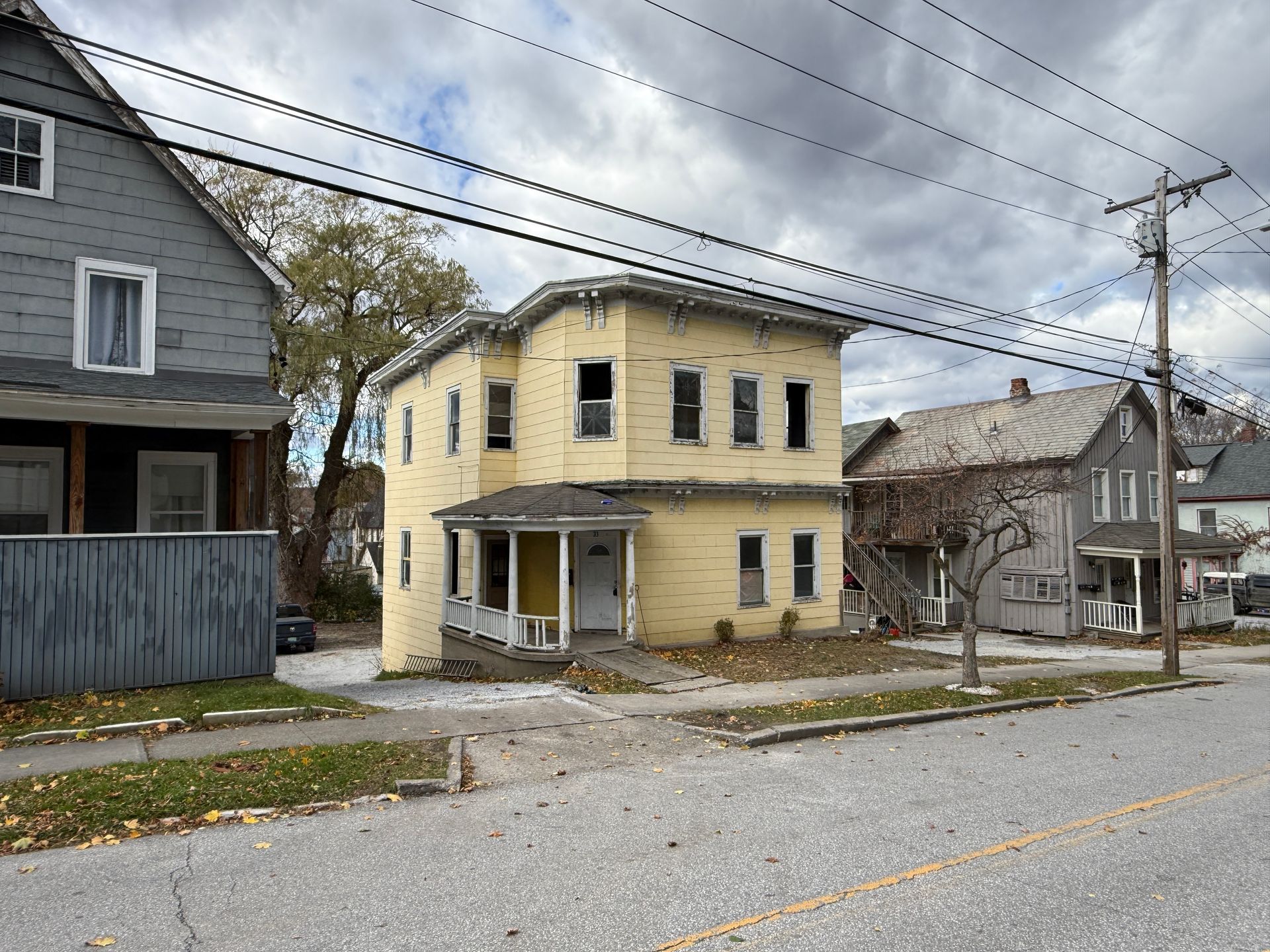 Yellow two-story house sits on a street, flanked by other houses under a cloudy sky.