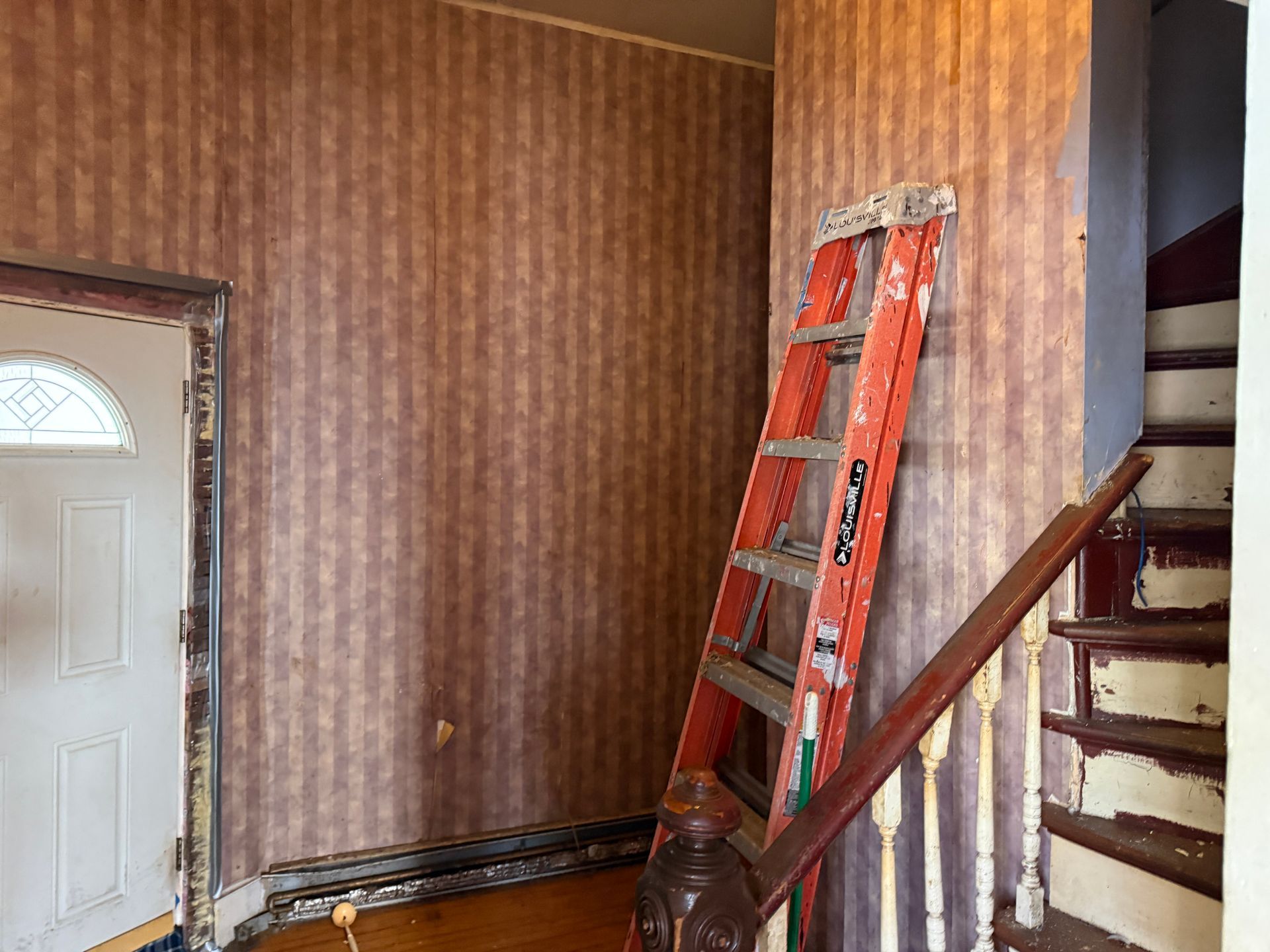 Interior hallway with a ladder leaning against a wall with striped wallpaper, stairs on the right, and a door on the left.