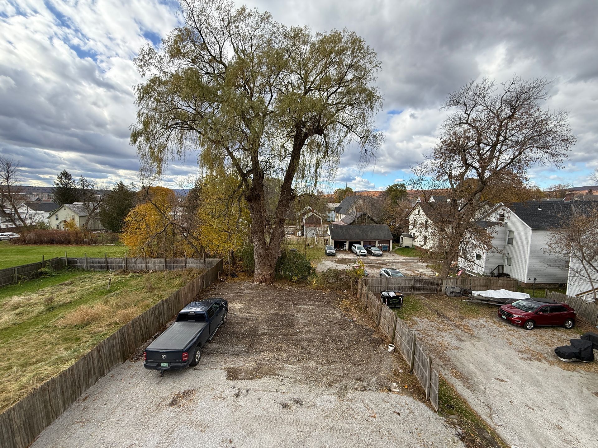 View of a gravel lot with a pickup truck, tree, and surrounding houses under a cloudy sky.