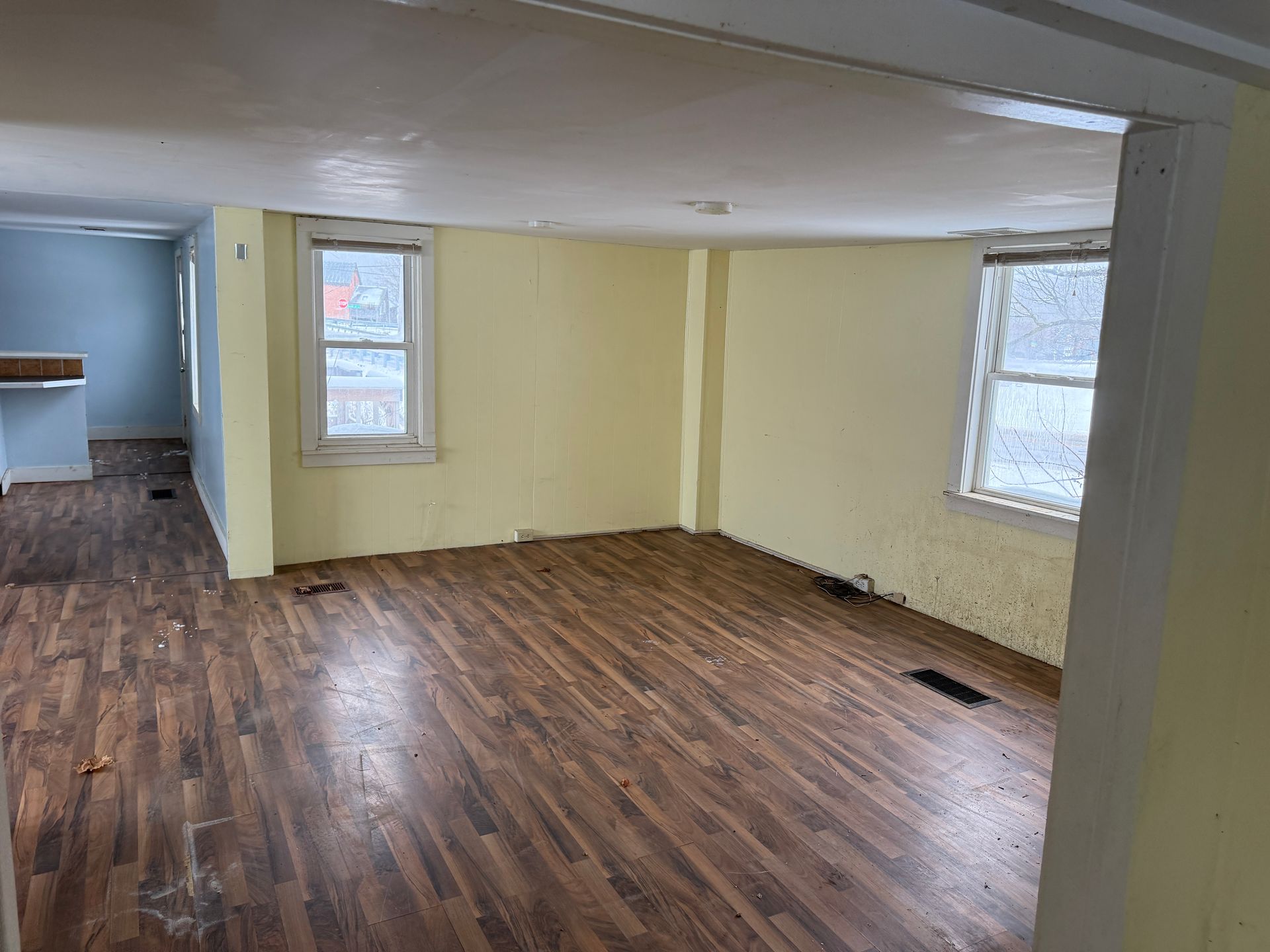 Empty living room with wood-look flooring, yellow walls, and two windows. A blue room is visible in the background.