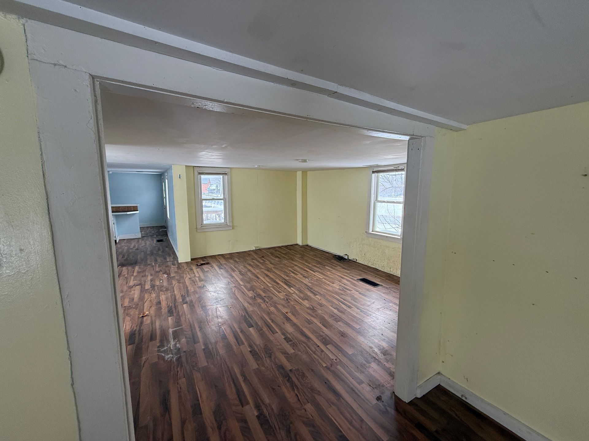 Interior view of a room with yellow walls, dark wood floor, and two windows. Another room is visible in the background.