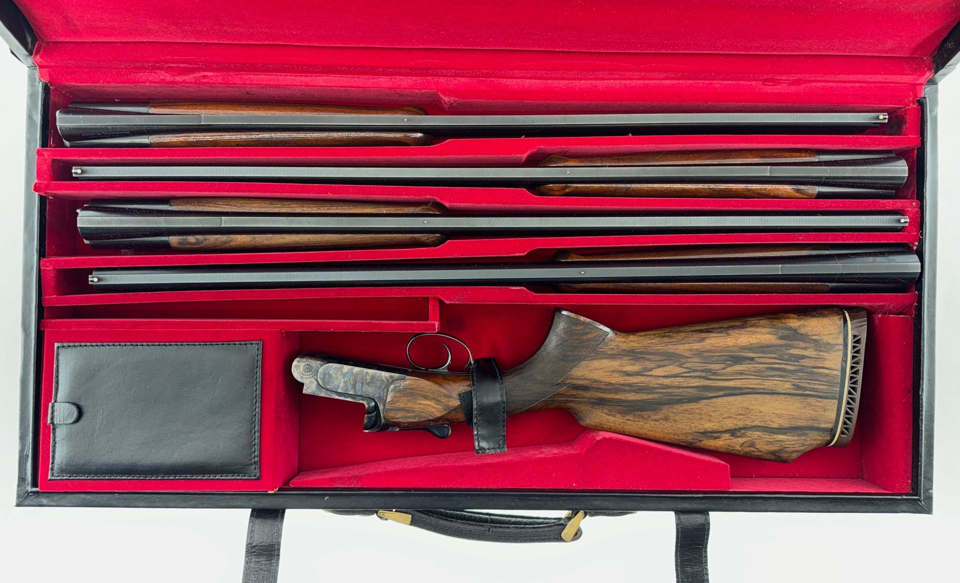 Gun parts organized in a red velvet-lined case, featuring several barrels and a wooden stock.
